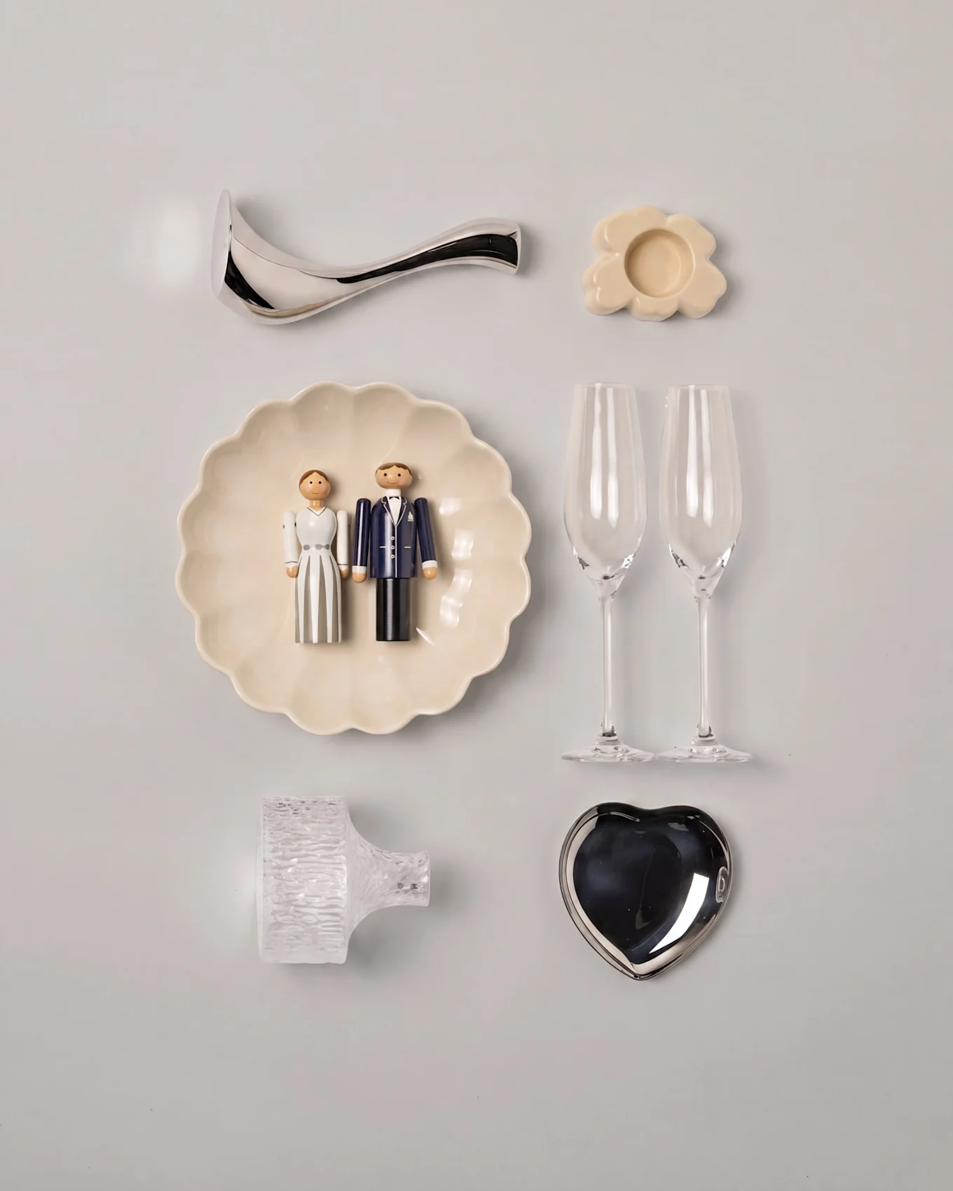 Disposición plana de boda con figuras de madera de novios en un plato festoneado, dos copas de champán, un plato plateado en forma de corazón y decoraciones variadas.