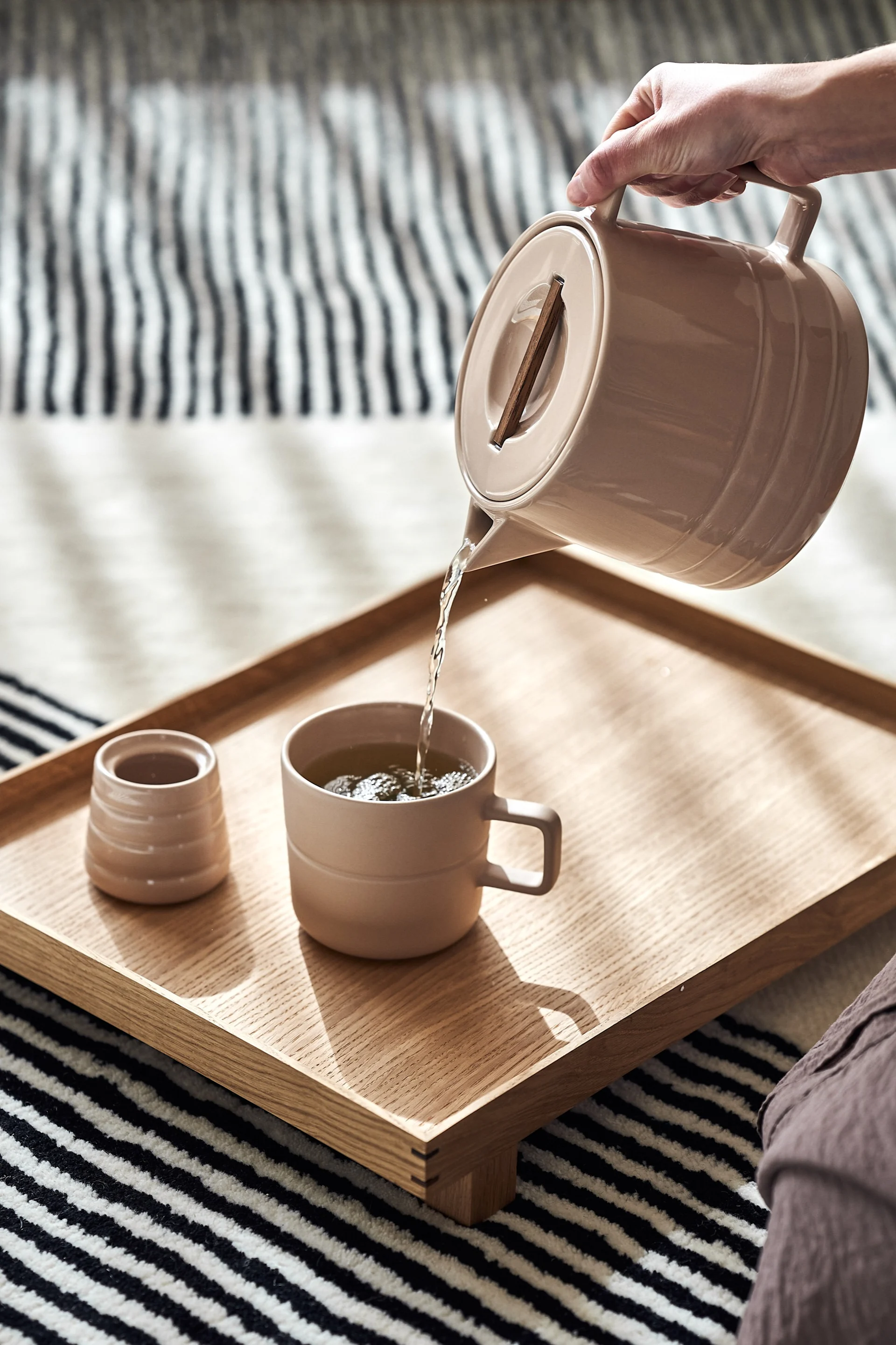 Mano sirviendo té de una tetera beige en una taza a juego sobre una bandeja de madera y una alfombra a rayas.