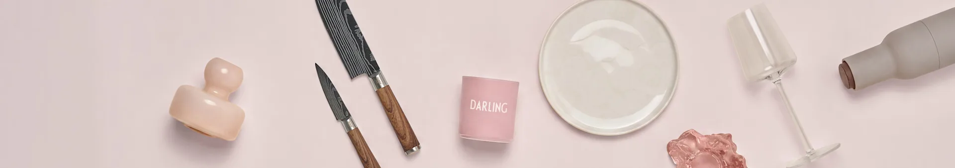 Surtido de artículos de cocina y comedor sobre fondo rosa: una escultura rosa, dos cuchillos estampados, un portavelas 'DARLING', un plato crema, una copa de vino transparente y una botella gris.