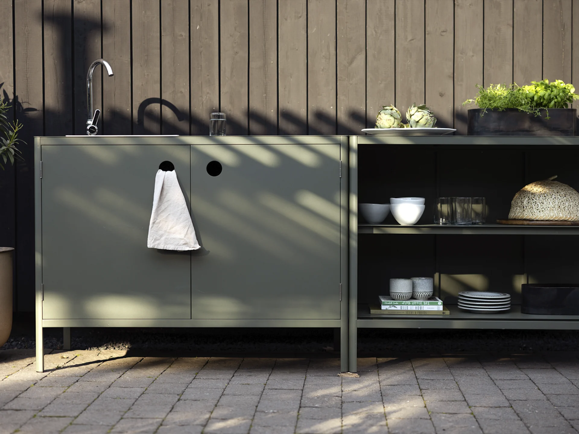 Cocina exterior con armarios verde oscuro, fregadero, grifo y estantes con alcachofas, hierbas y vajilla.