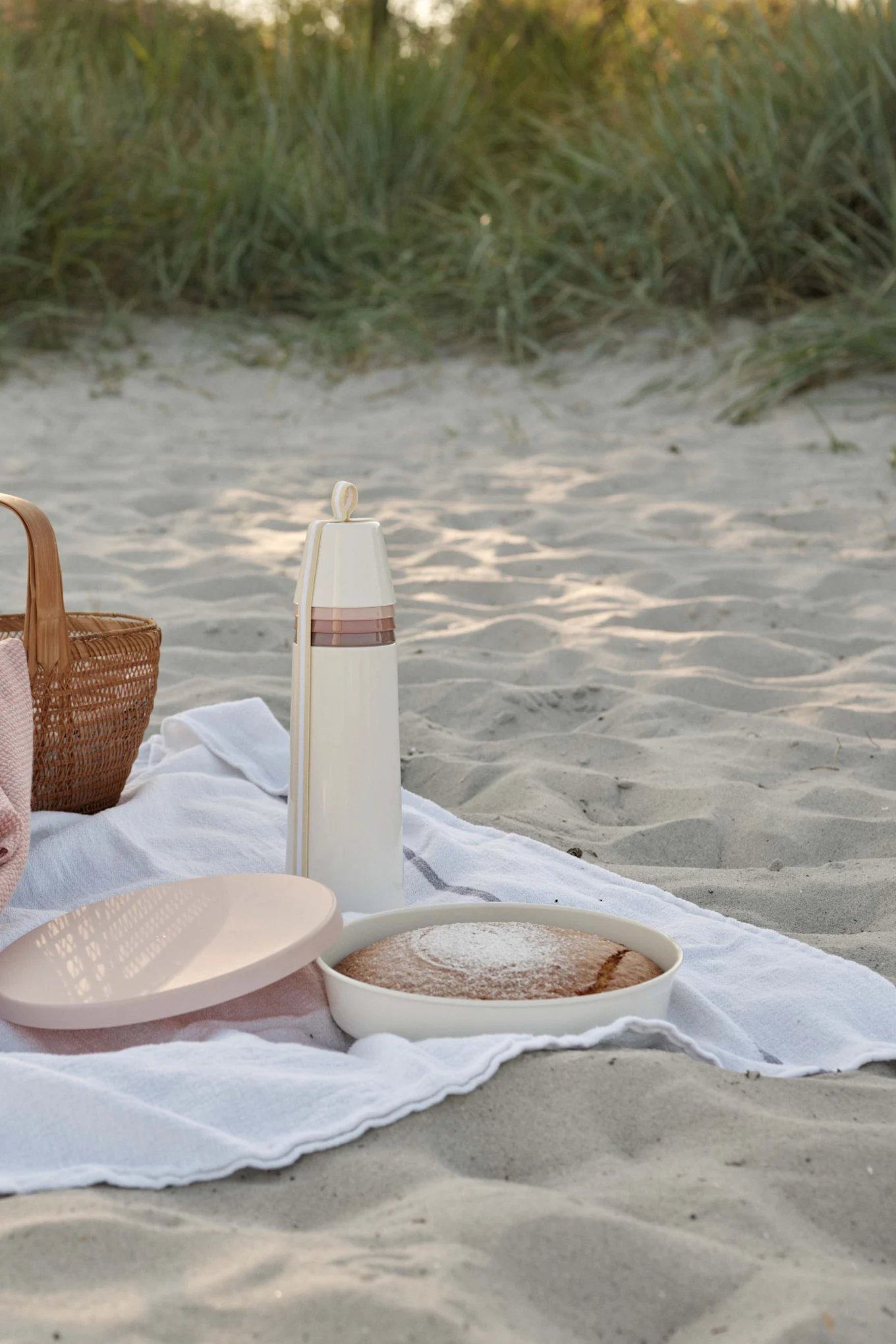Picnic en una playa de arena con un termo crema, pastel con azúcar glas y una cesta de mimbre.