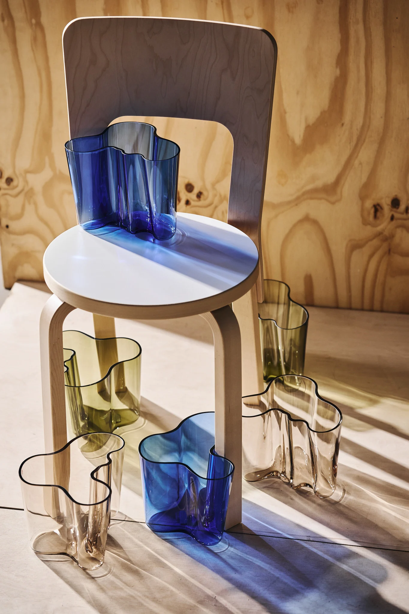 Múltiples jarrones de cristal ondulados en azul, verde oliva y transparente, dispuestos sobre y alrededor de un taburete de madera claro con una silla de madera al fondo. La luz del sol proyecta sombras de colores.