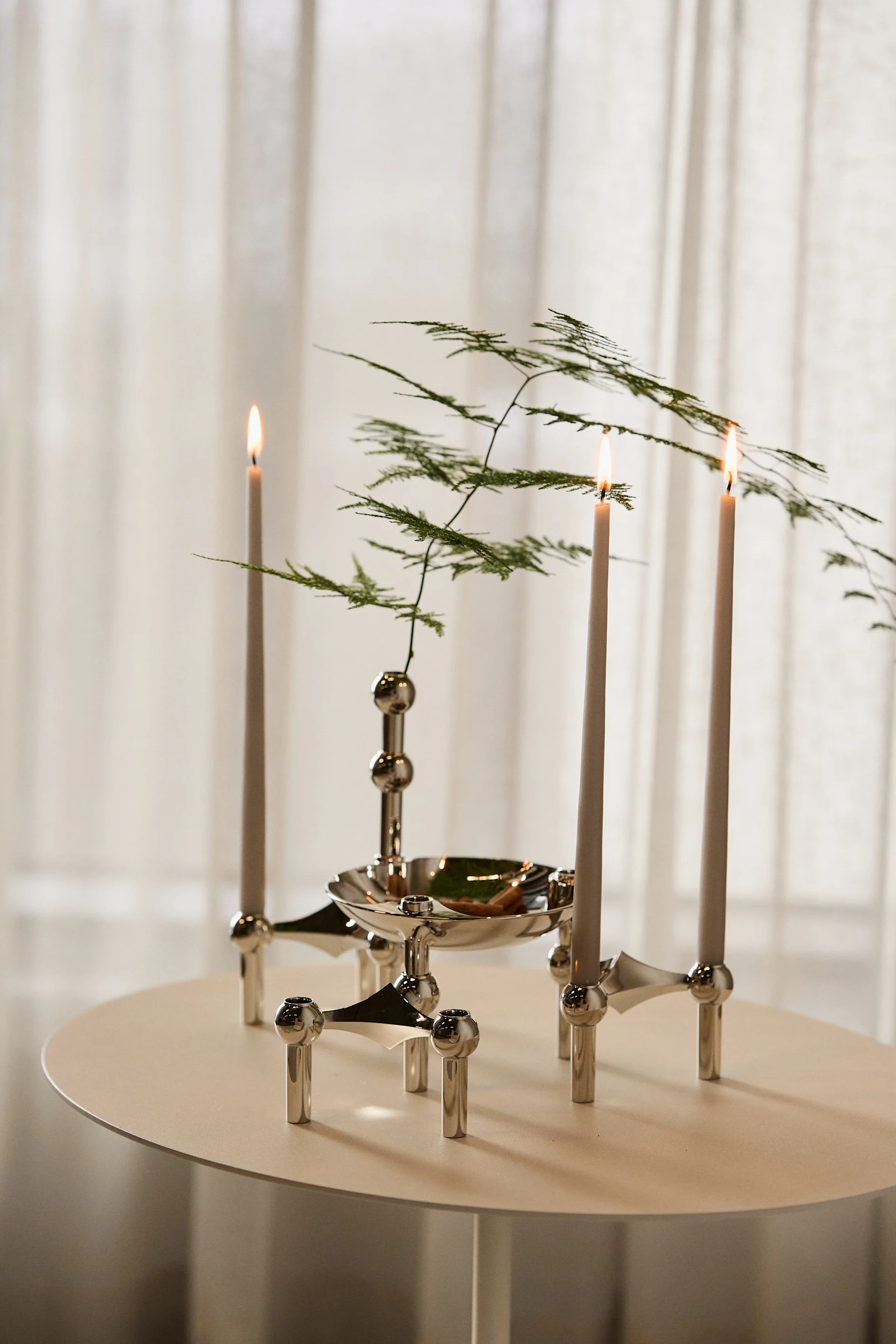 Tres velas encendidas en un candelabro modular de plata con follaje verde, sobre una mesa redonda clara.