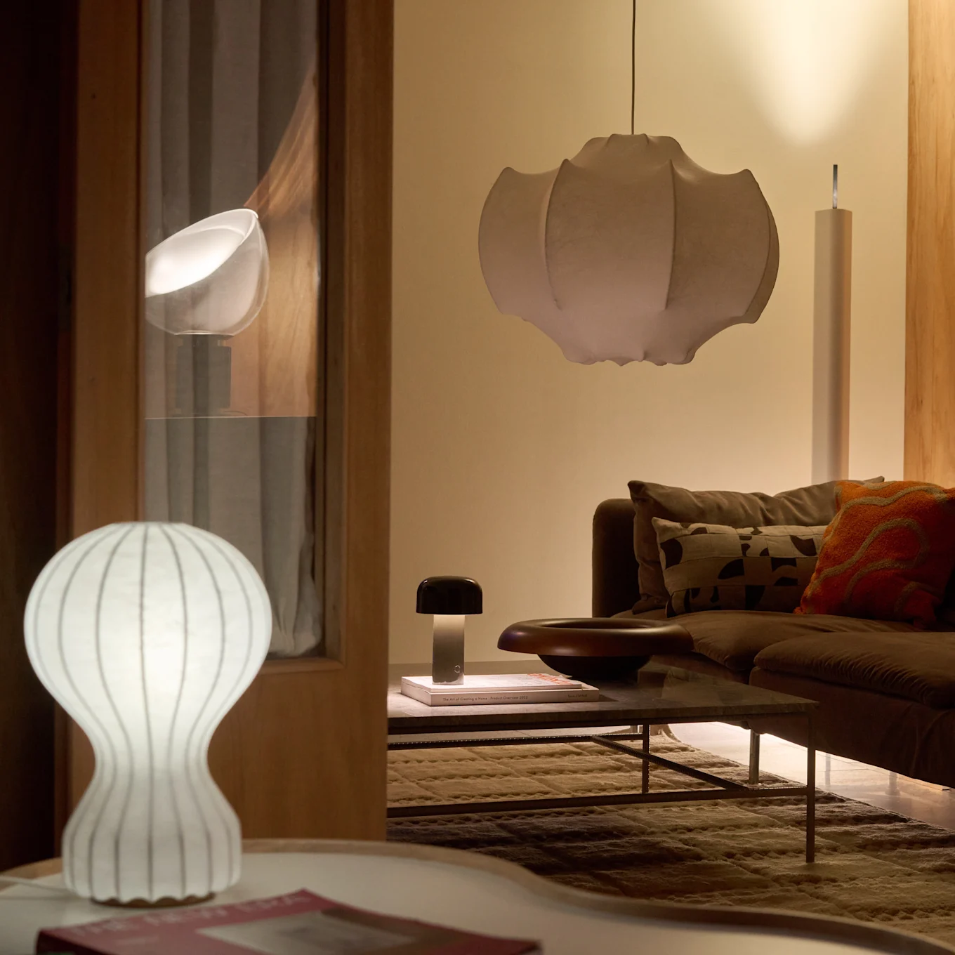 Un salón con sofá y mesa de centro marrones, iluminado por una lámpara de mesa blanca acanalada, una lámpara con forma de seta oscura y una gran lámpara colgante plisada, creando un brillo cálido.