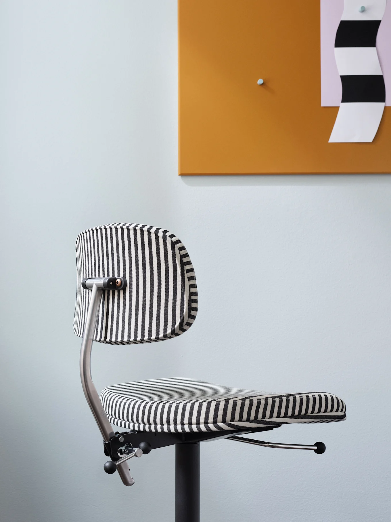 Una moderna silla de oficina con rayas verticales blancas y negras, colocada contra una pared azul claro y un tablero naranja.