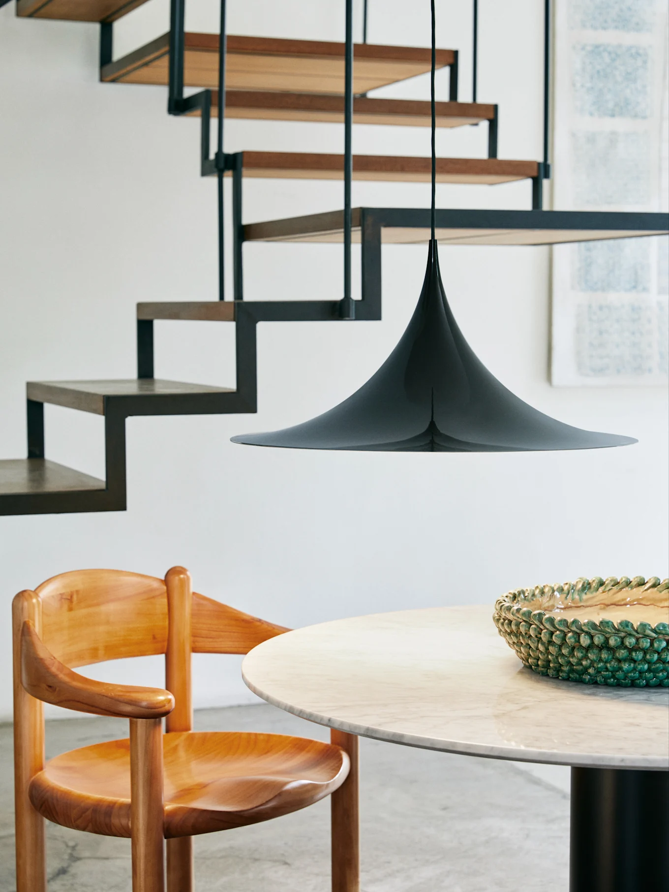 Una lámpara colgante negra cónica cuelga sobre una mesa redonda de mármol y una silla de madera, con una escalera moderna al fondo.