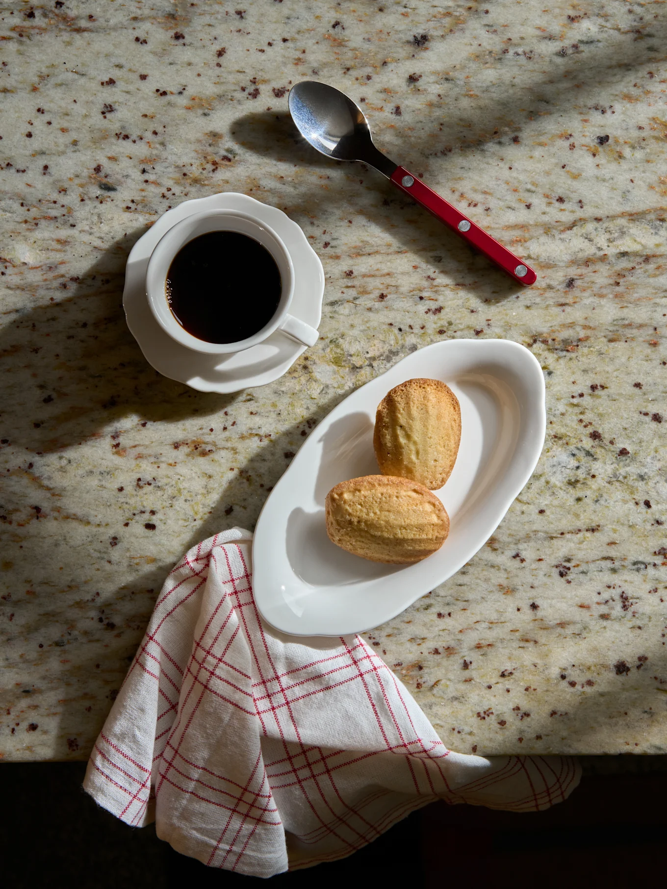 Vista superior de café negro en taza blanca, dos madeleines en plato blanco, cuchara y servilleta a rayas sobre mármol.