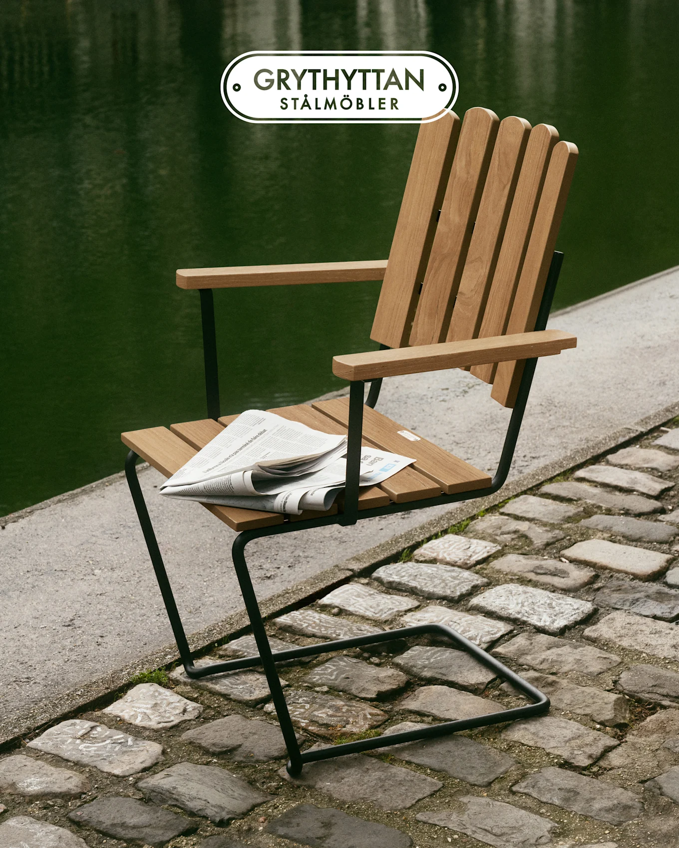 Silla de lamas de madera con estructura de metal negro en camino empedrado junto a agua verde, periódico en el asiento.