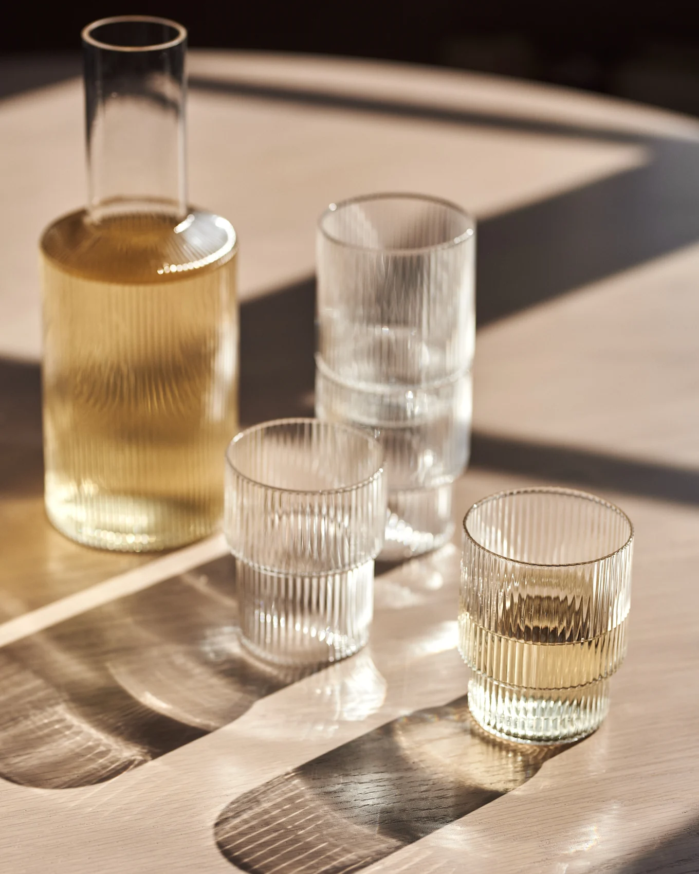 Garrafa de vidrio y vasos acanalados en una mesa de madera con luz solar.