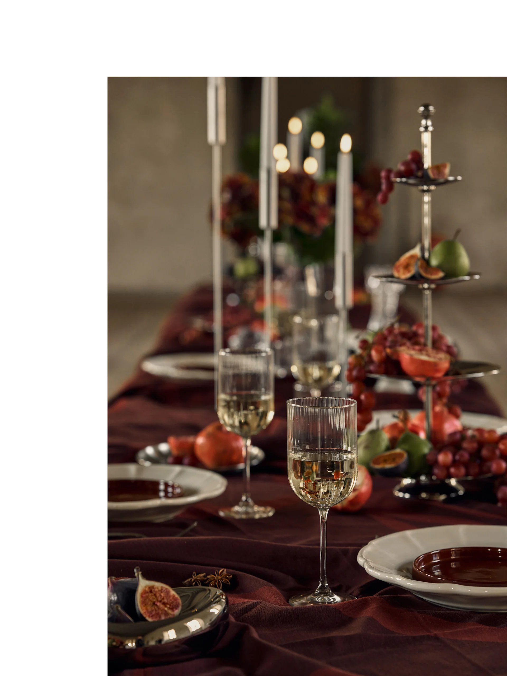 Mesa festiva puesta con copas de vino blanco, platos y un expositor escalonado con frutas coloridas y luz de velas.