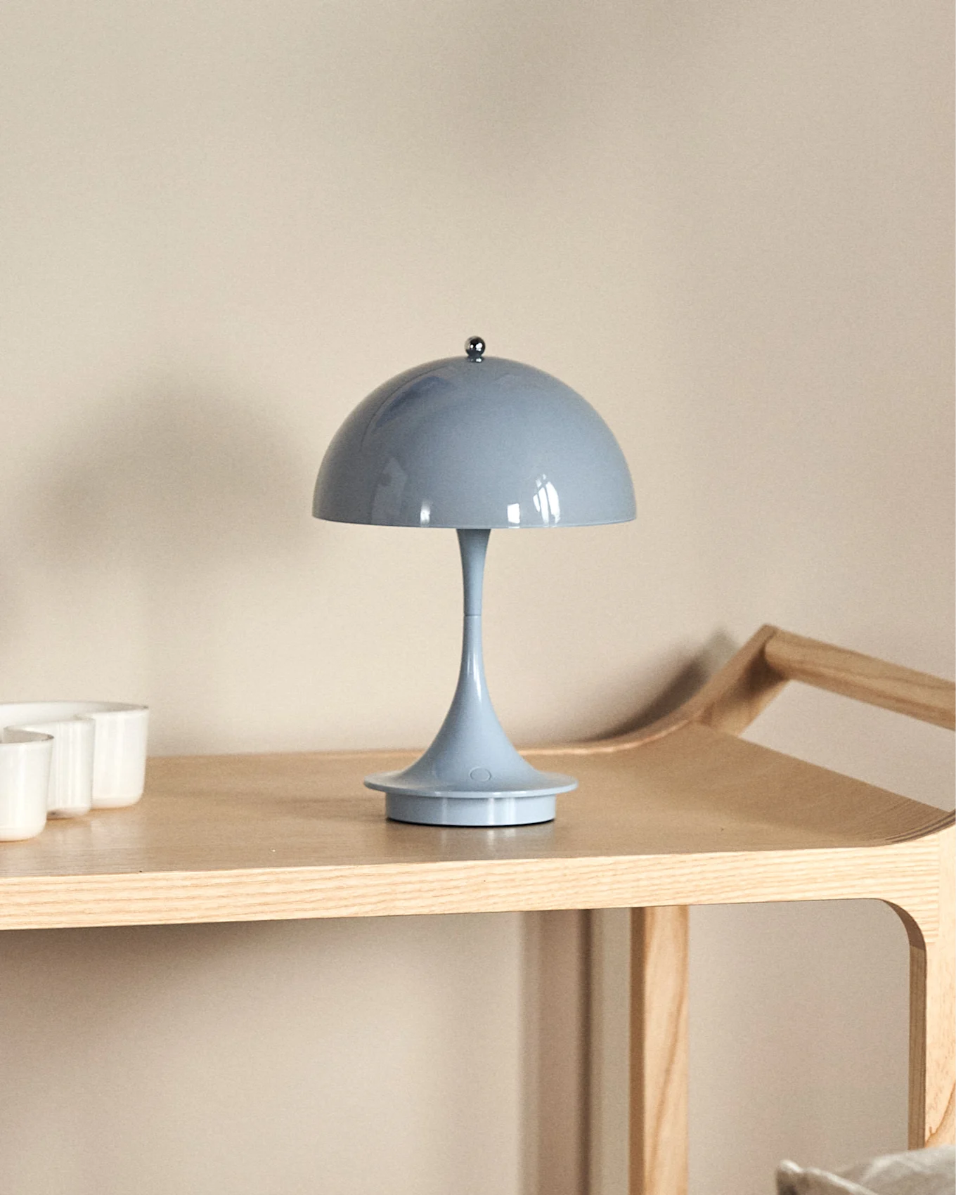 Una lámpara de mesa azul claro con forma de hongo sobre un estante de madera clara, con tazas blancas parcialmente visibles a la izquierda.