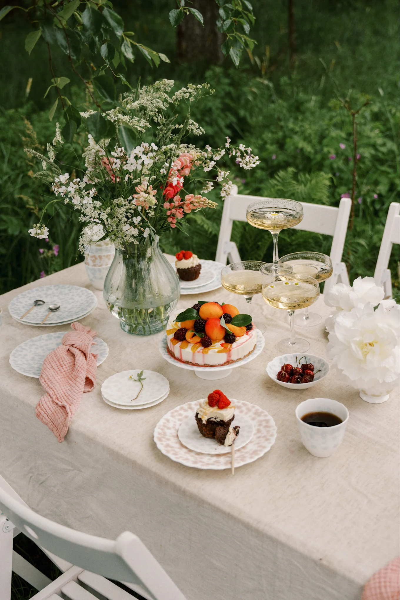 Mesa al aire libre con jarrón de flores, champán, pastel de frutas y cupcakes con bayas.