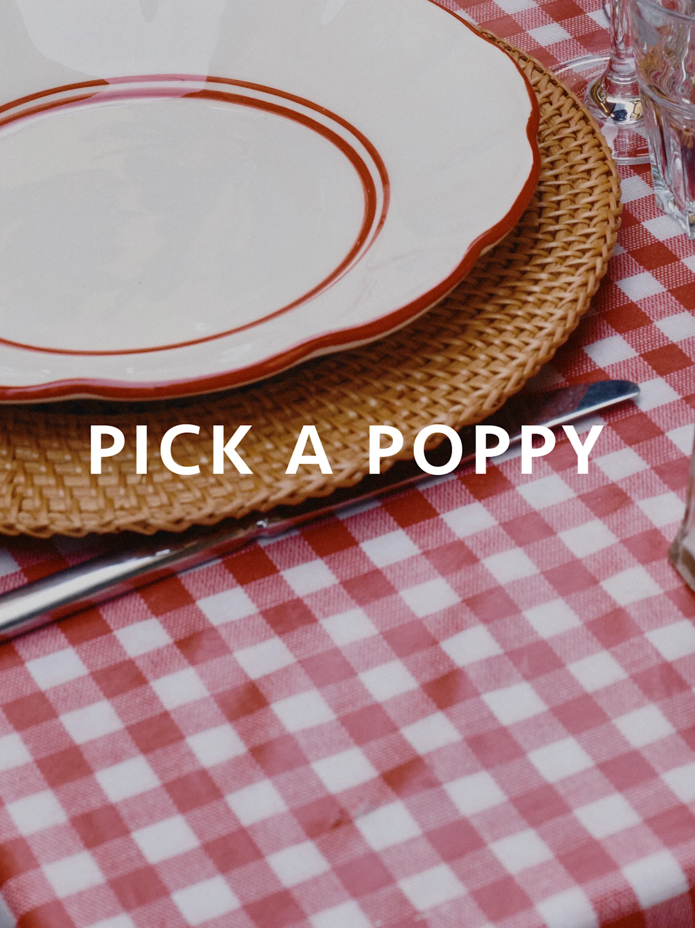 Mesa puesta con mantel a cuadros rojo y blanco, salvamantel de mimbre y plato blanco con borde rojo. Texto: PICK A POPPY.