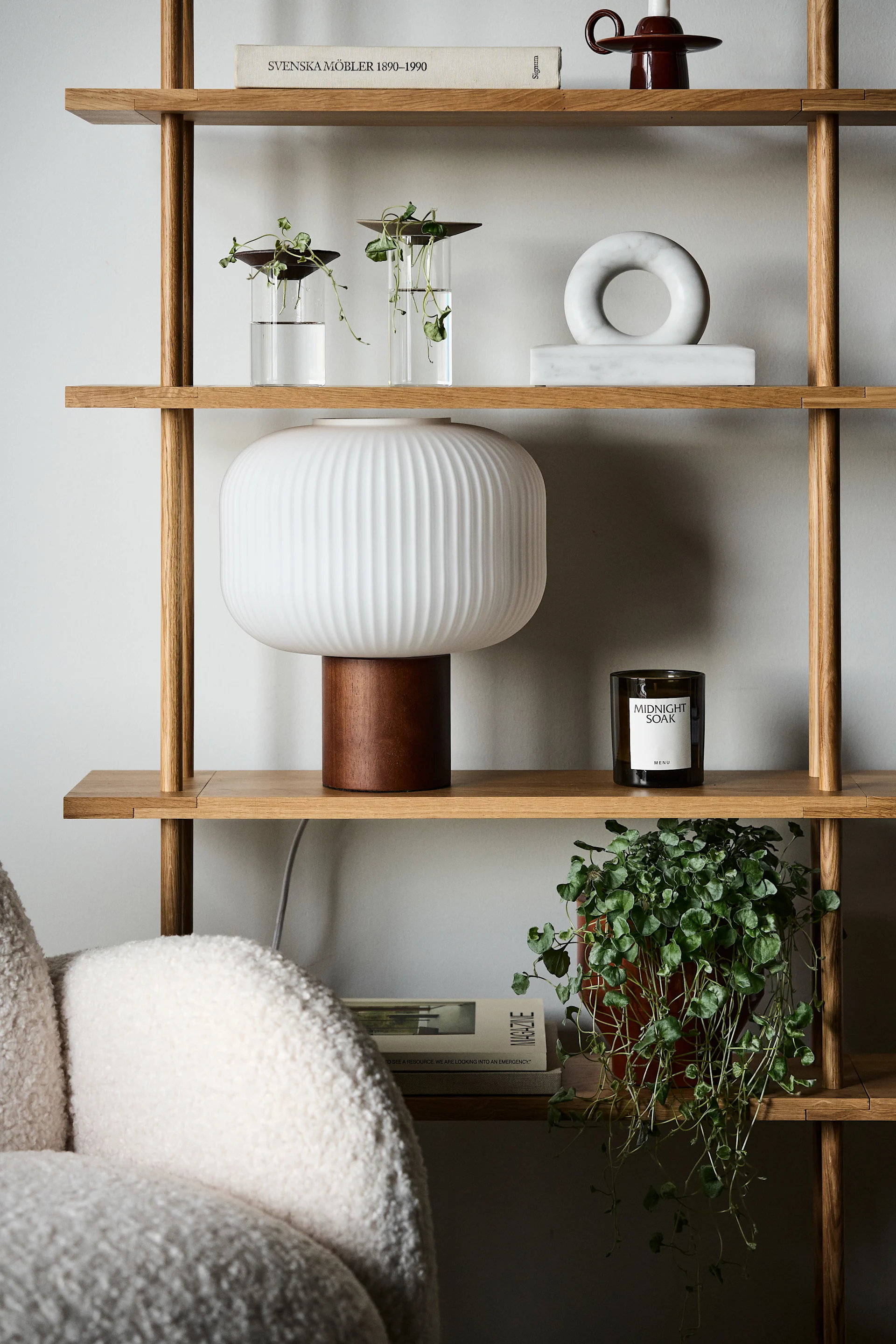 Una estantería de madera clara con plantas, una lámpara blanca estriada, decoración de mármol, libro y vela.