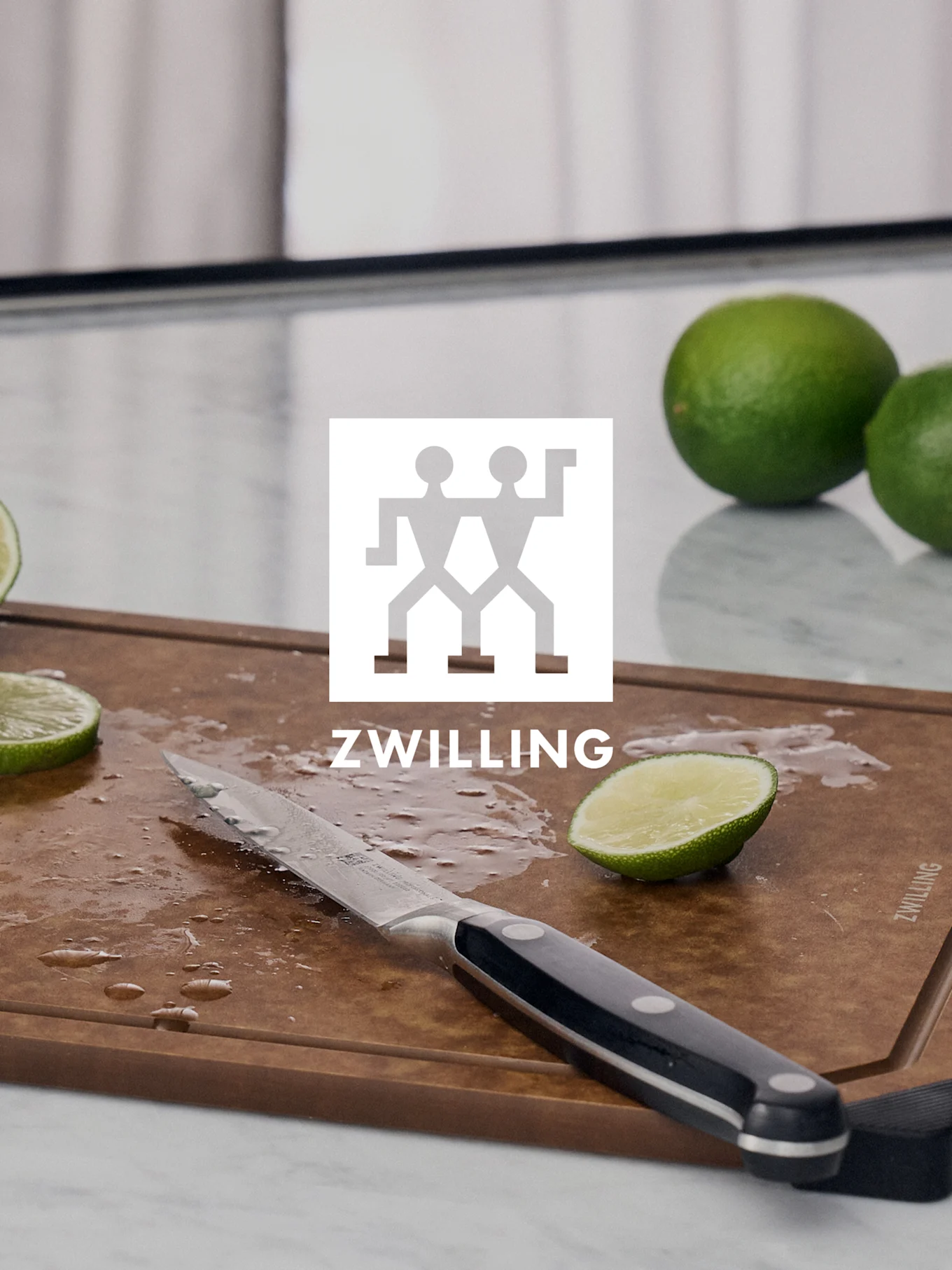 Logo de ZWILLING sobre una tabla de cortar con un cuchillo, lima cortada y limas enteras.