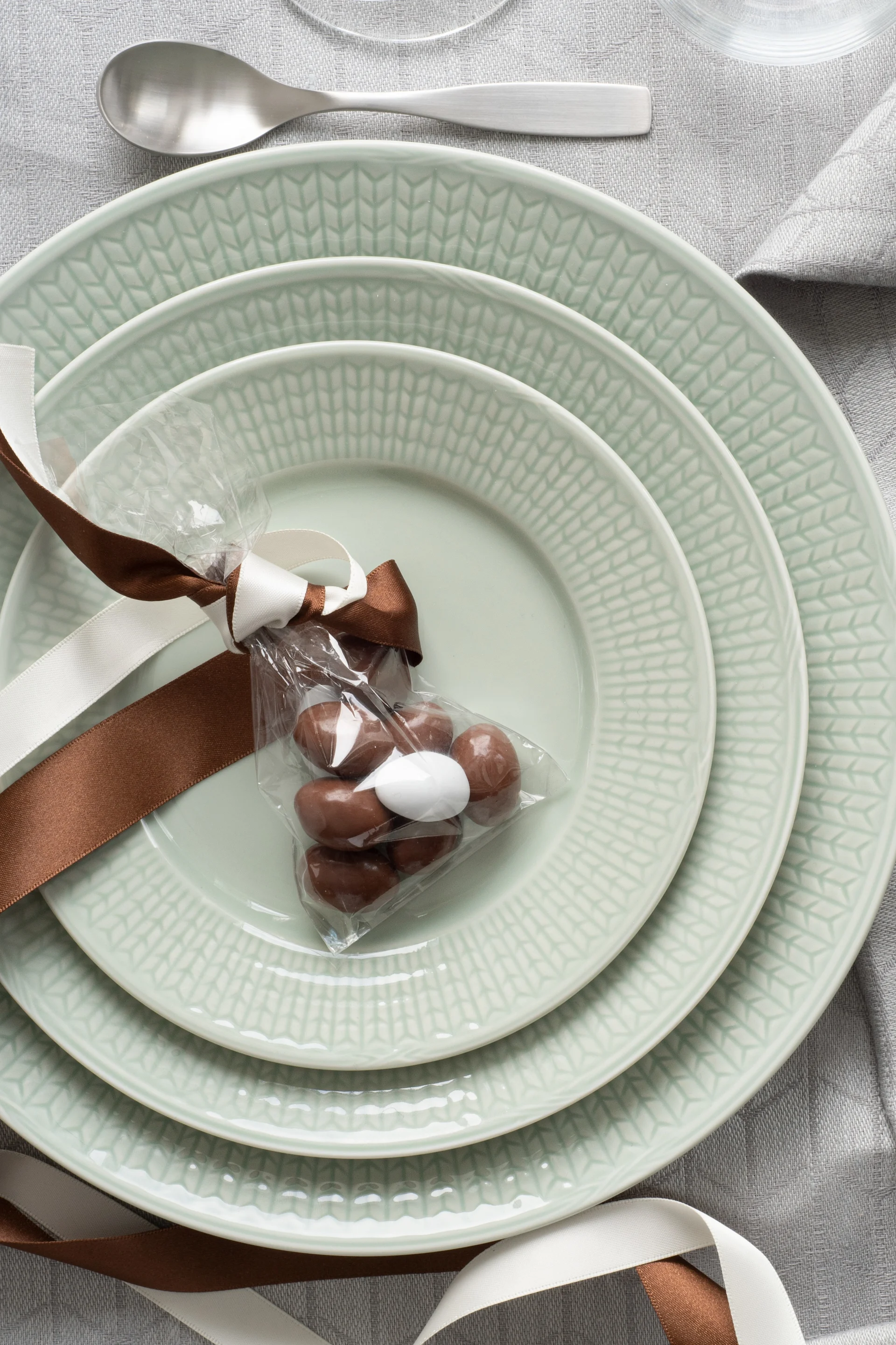 Platos apilados de color verde claro con una bolsa de regalo de chocolates sobre un mantel gris, junto a cubiertos plateados.