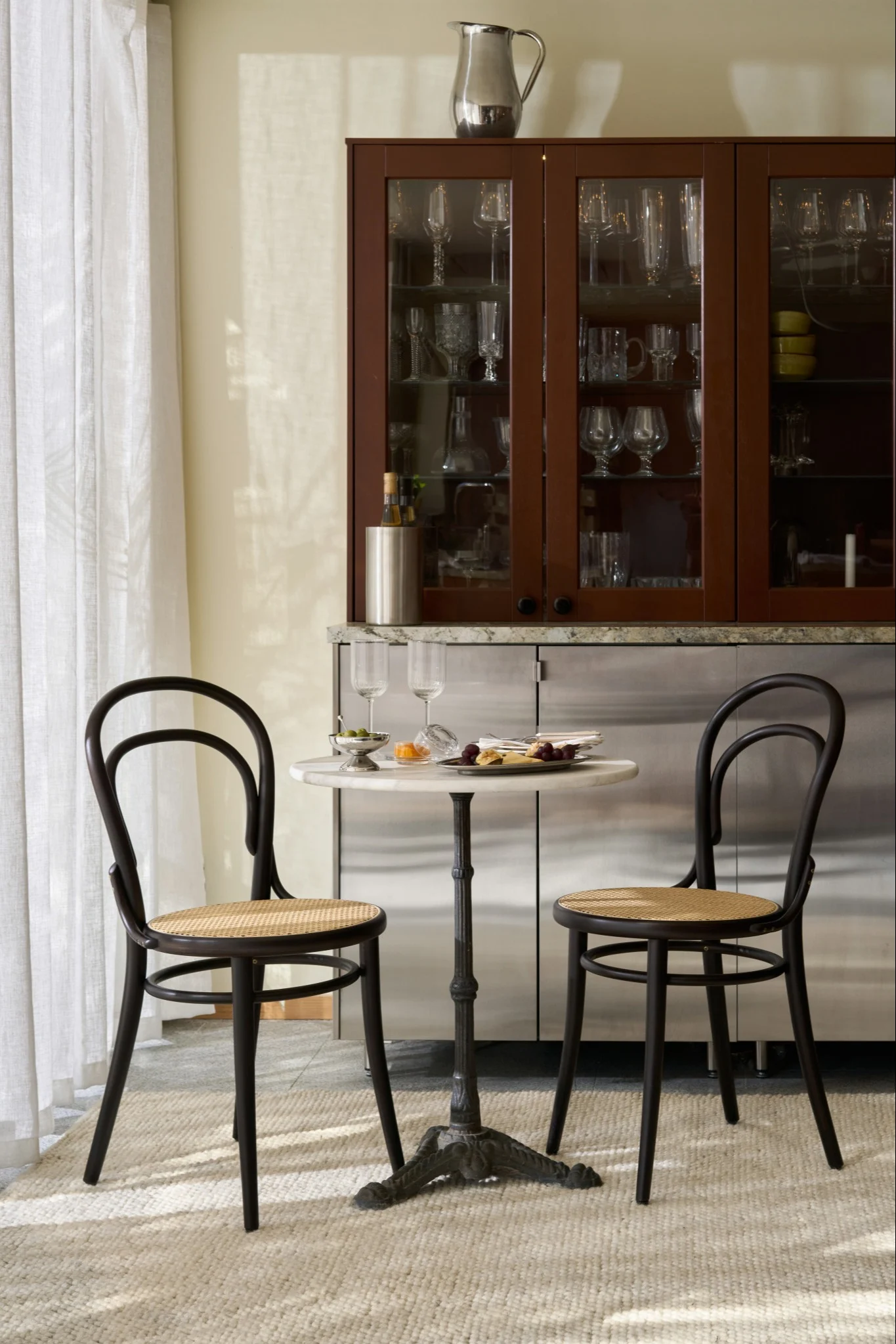 Dos sillas oscuras de madera curvada y una pequeña mesa de mármol con aperitivos frente a un mueble bar con cristalería.