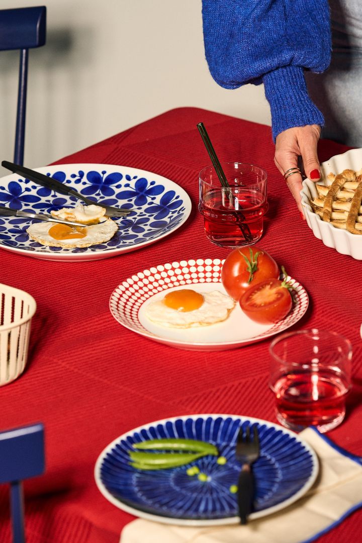 Las tendencias de color para interiores de 2023 incluyen el contraste de rojo y azul. Aquí puedes ver una mesa puesta con platos azules Mon Amie de Rörstrand sueco y un mantel rojo de NJRD.
