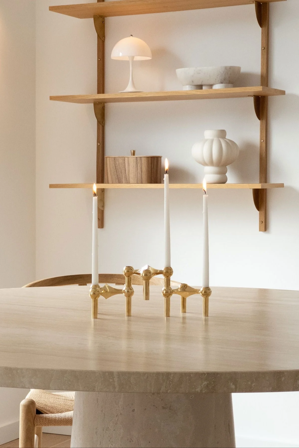 Un candelabro dorado con tres velas encendidas sobre una mesa redonda, con estantes con objetos decorativos detrás.