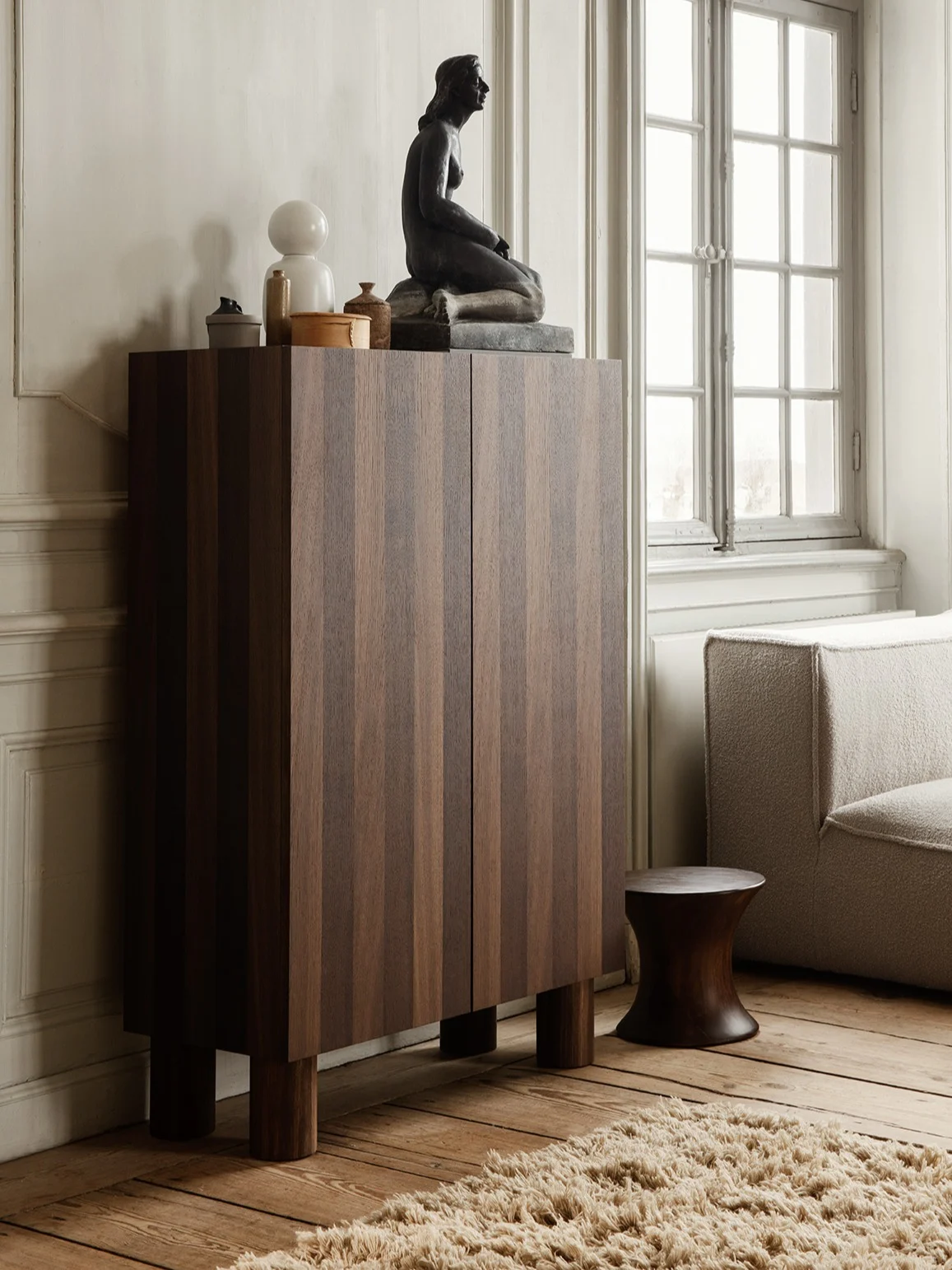 Armario de madera oscura con rayas verticales, coronado por una escultura sentada, lámpara blanca y decoración, junto a un sofá y taburete oscuro.