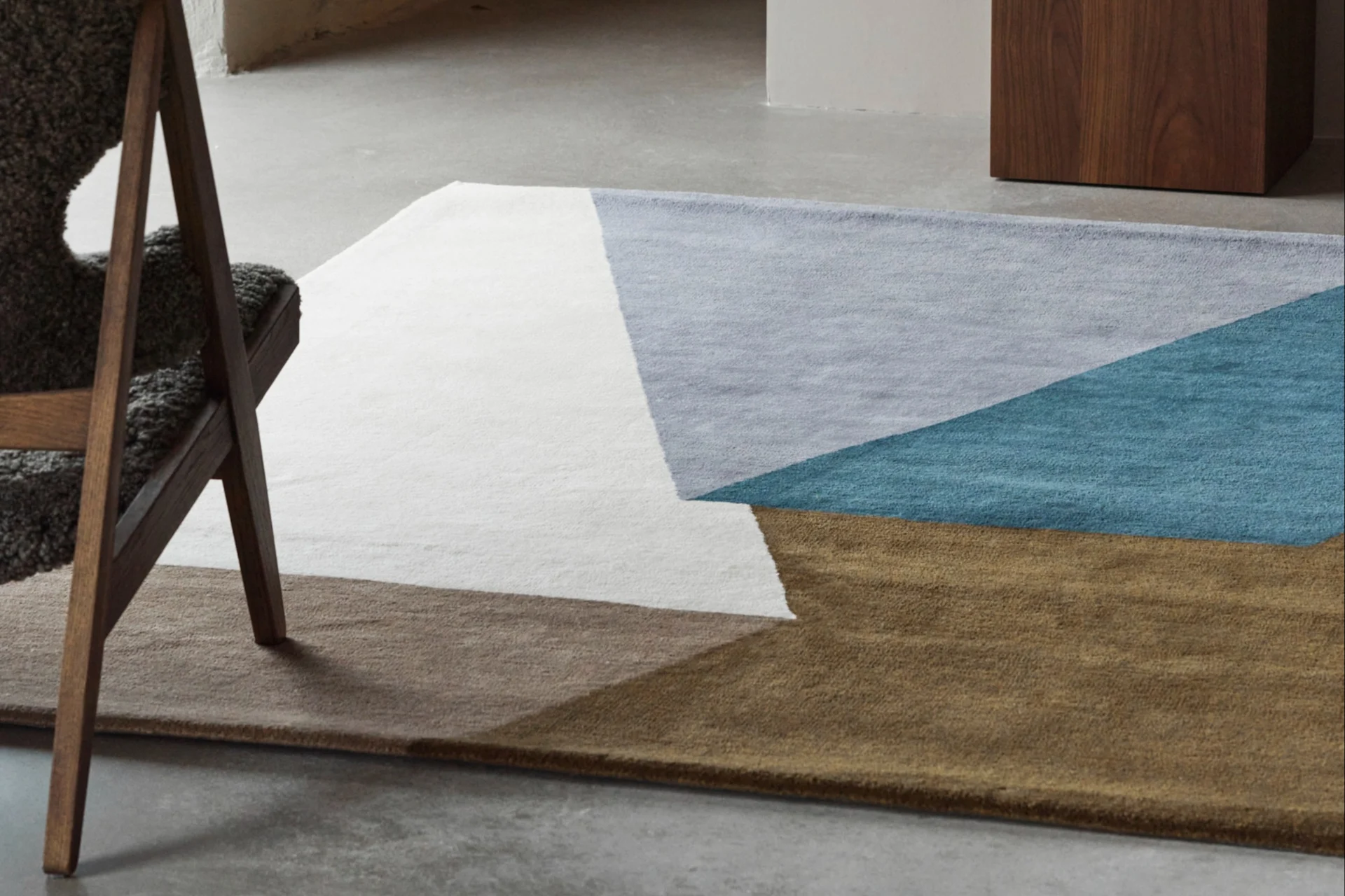 Una alfombra geométrica en blanco, azul claro, turquesa y marrón sobre un suelo de hormigón, con una silla de madera y un jarrón azul.