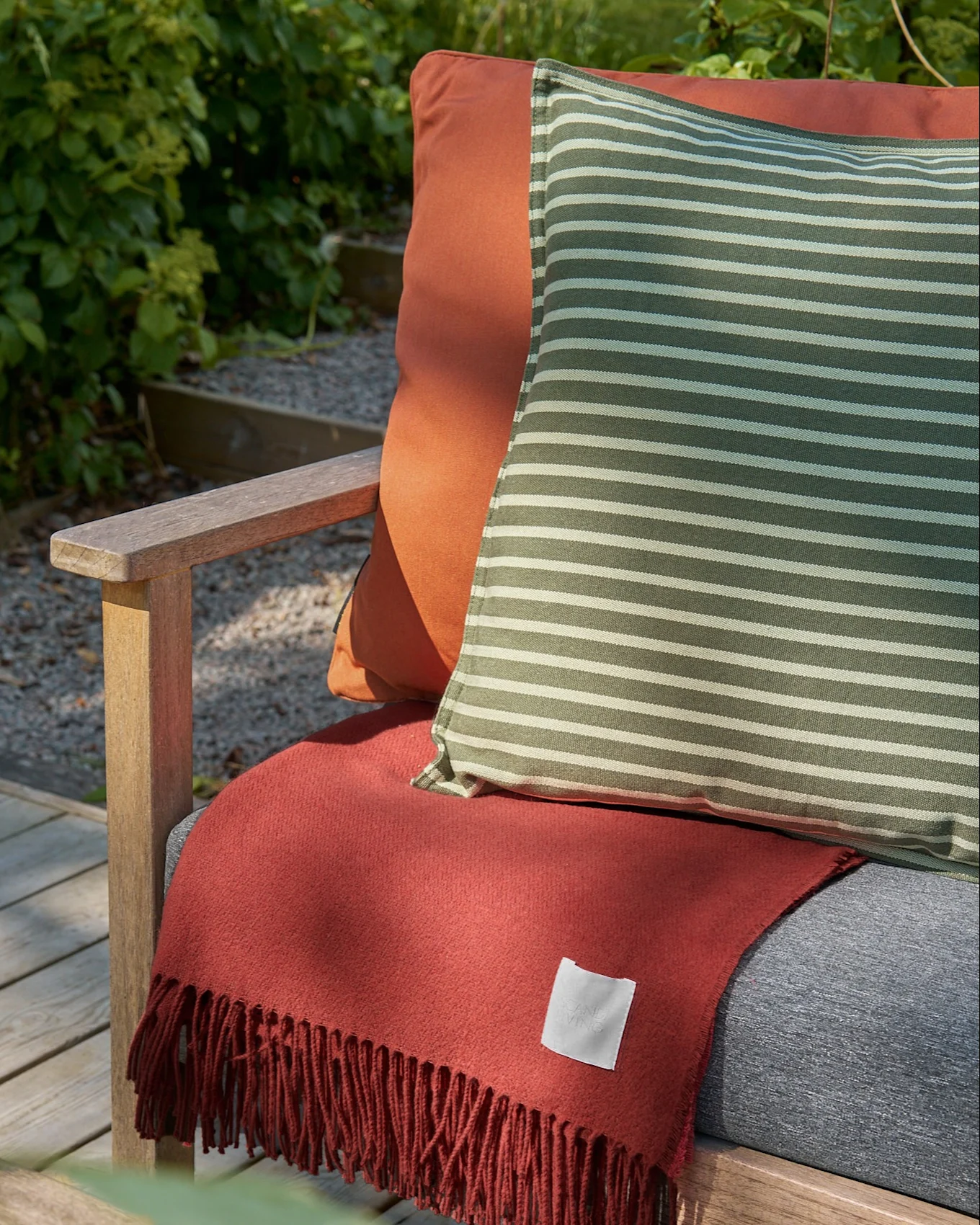 Silla de madera para exterior con cojín naranja, almohada a rayas verdes y blancas, y manta de flecos color terracota.