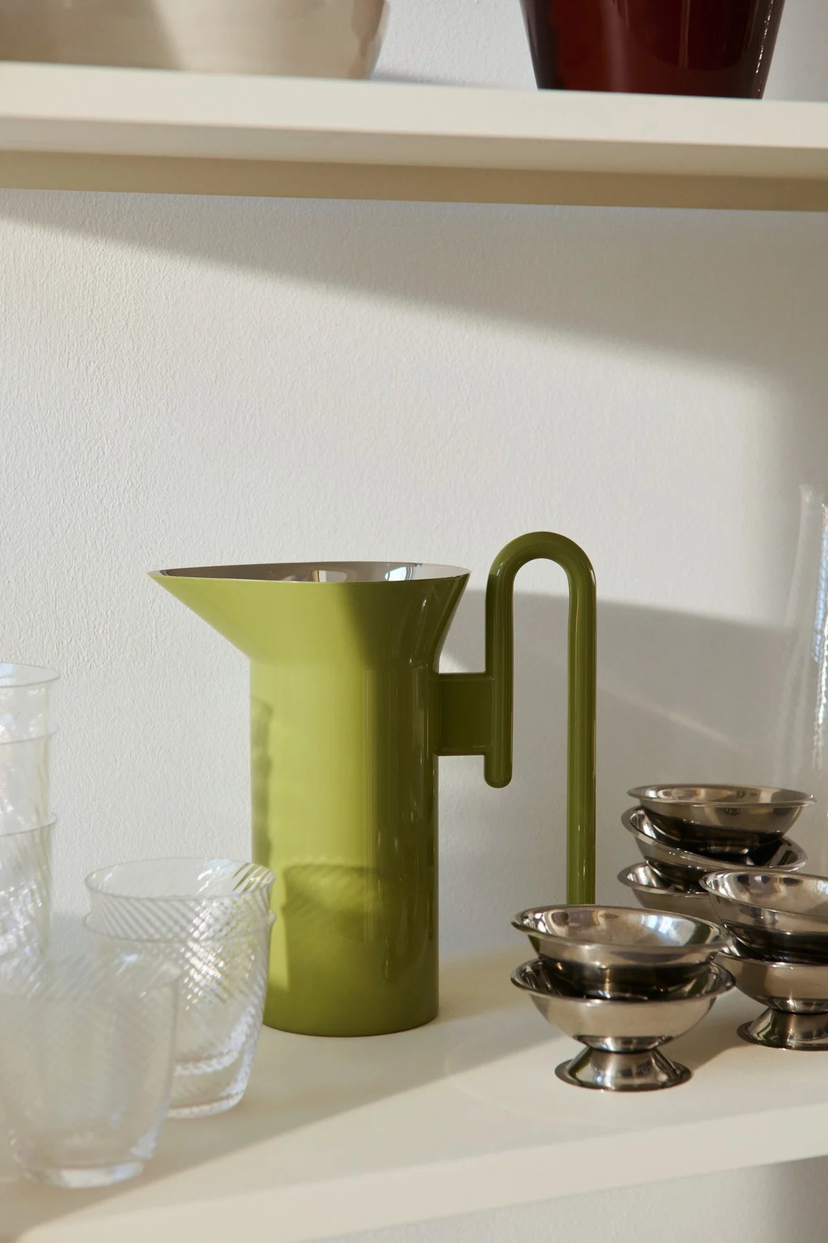 Una jarra verde claro con asa única en forma de U, vasos transparentes texturizados y pequeños cuencos plateados en una estantería.