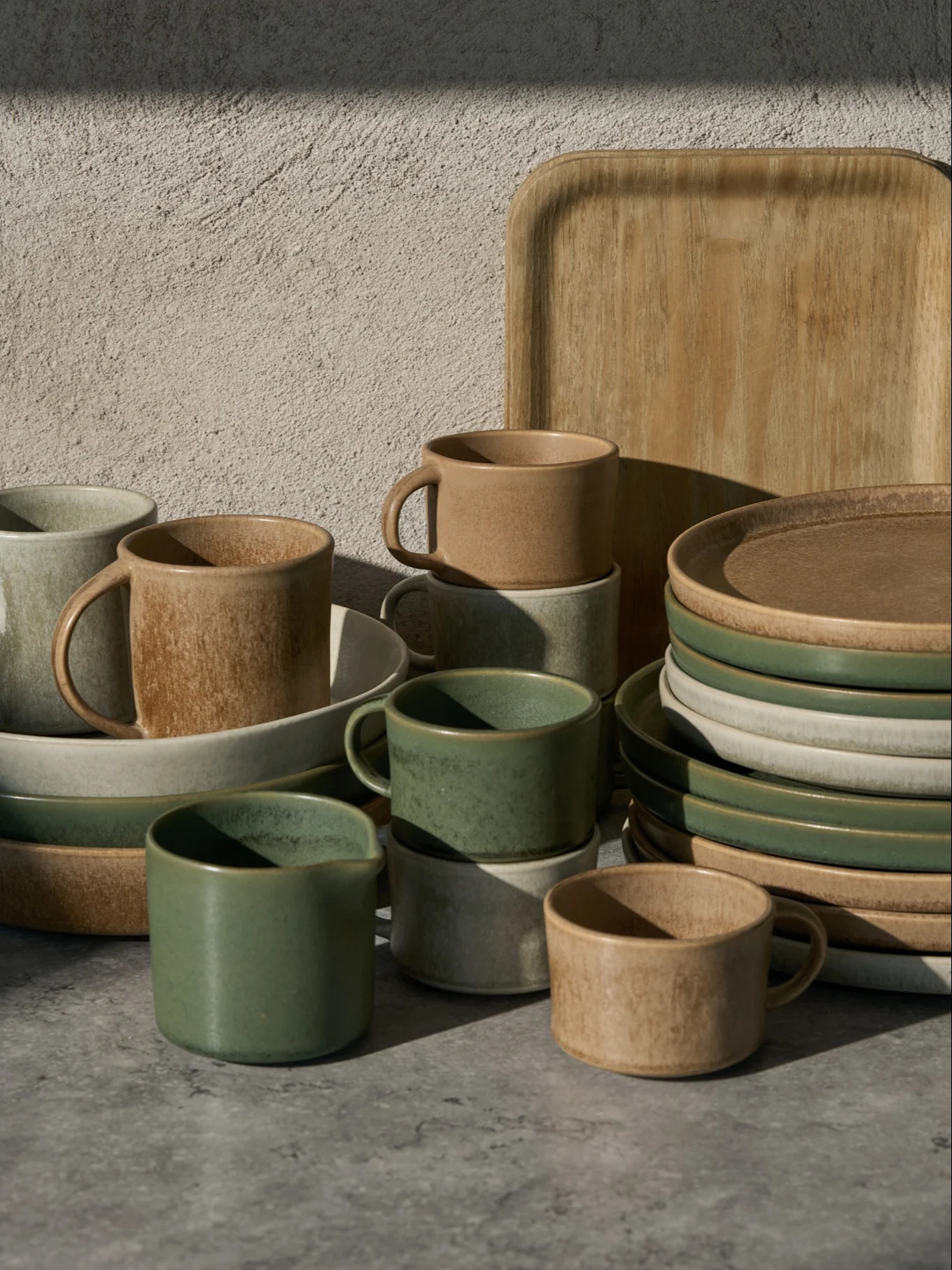 Colección de tazas, cuencos y platos de cerámica verdes, beige y gris claro, con una bandeja de madera, sobre una superficie gris.