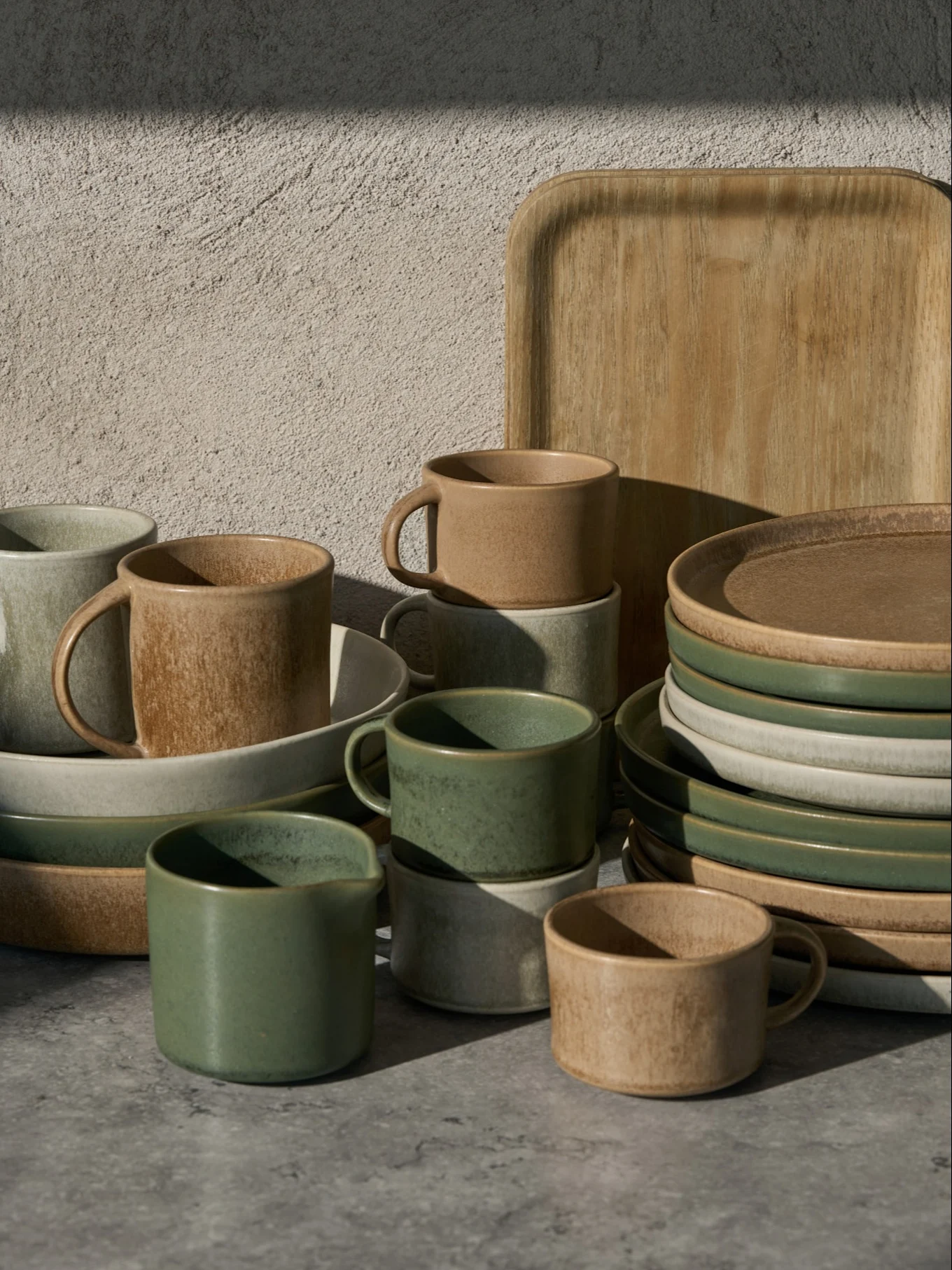 Colección de tazas, cuencos y platos de cerámica verdes, beige y gris claro, con una bandeja de madera, sobre una superficie gris.