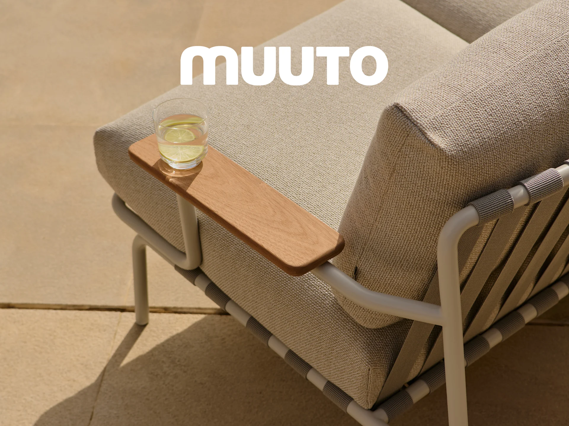 Primer plano de un sofá exterior con cojines de tela clara, un reposabrazos de madera con un vaso de agua y el logo blanco de Muuto.