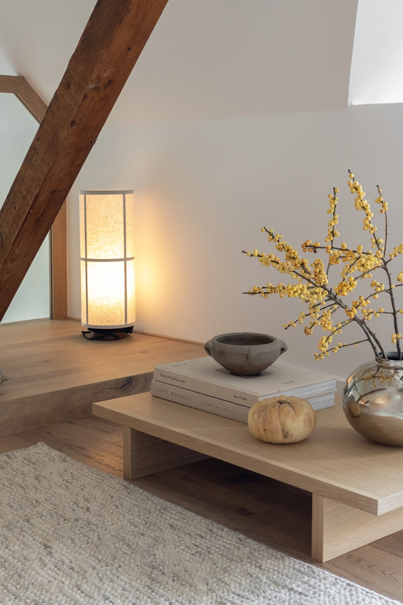 Sala de estar acogedora en el ático con lámpara alta, viga de madera, mesa de café baja con bayas amarillas, libros y calabaza.