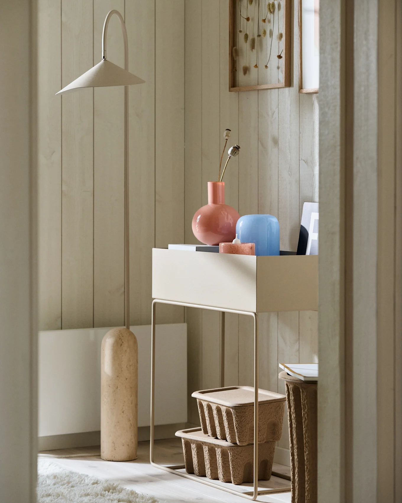 Rincón minimalista con una lámpara de pie beige alta, una mesa consola con jarrones rosa y azul, y cajas de almacenamiento marrones texturizadas.