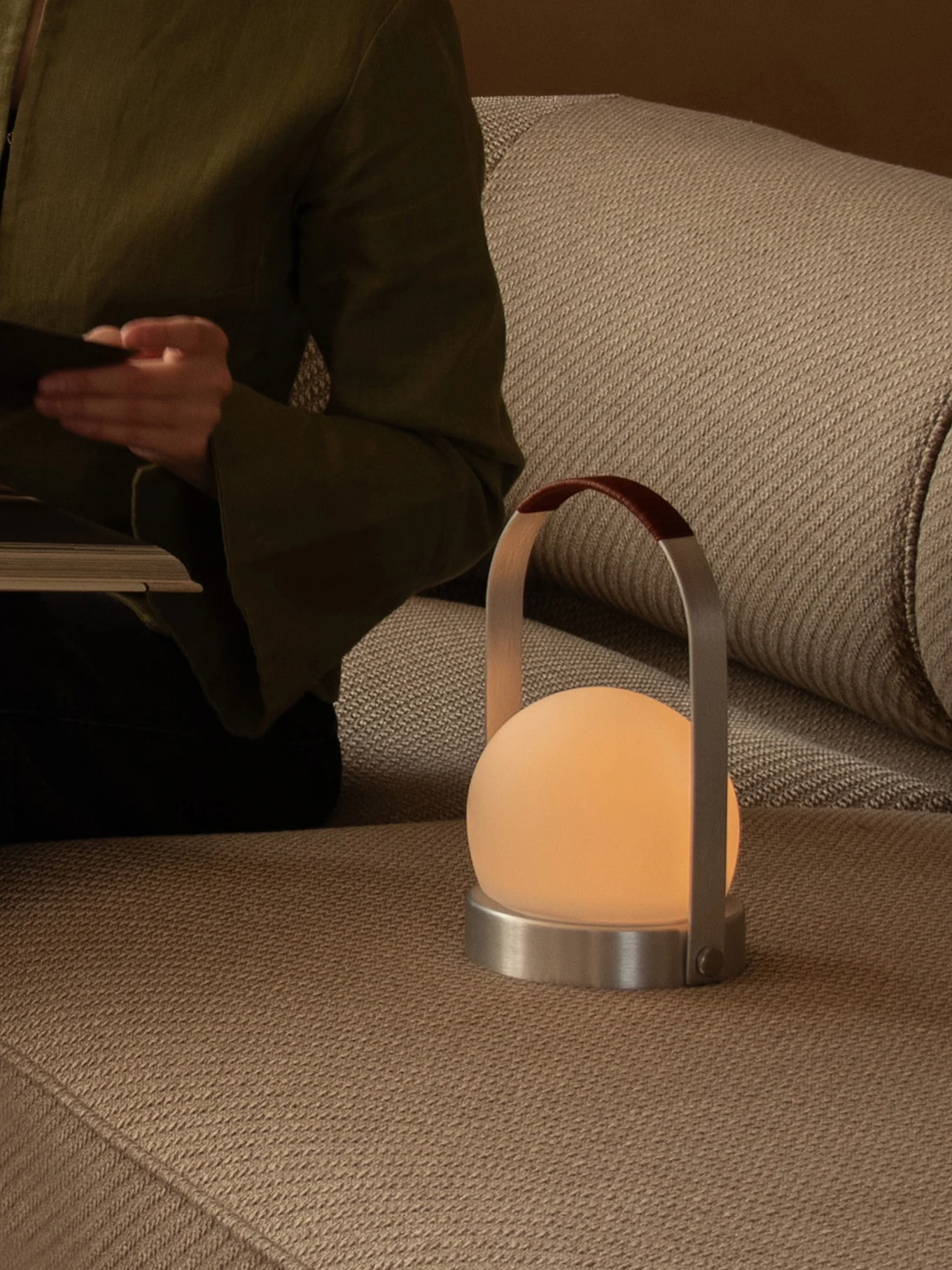 Persona leyendo en sofá con lámpara portátil encendida, con globo esmerilado, base de metal y asa de cuero.