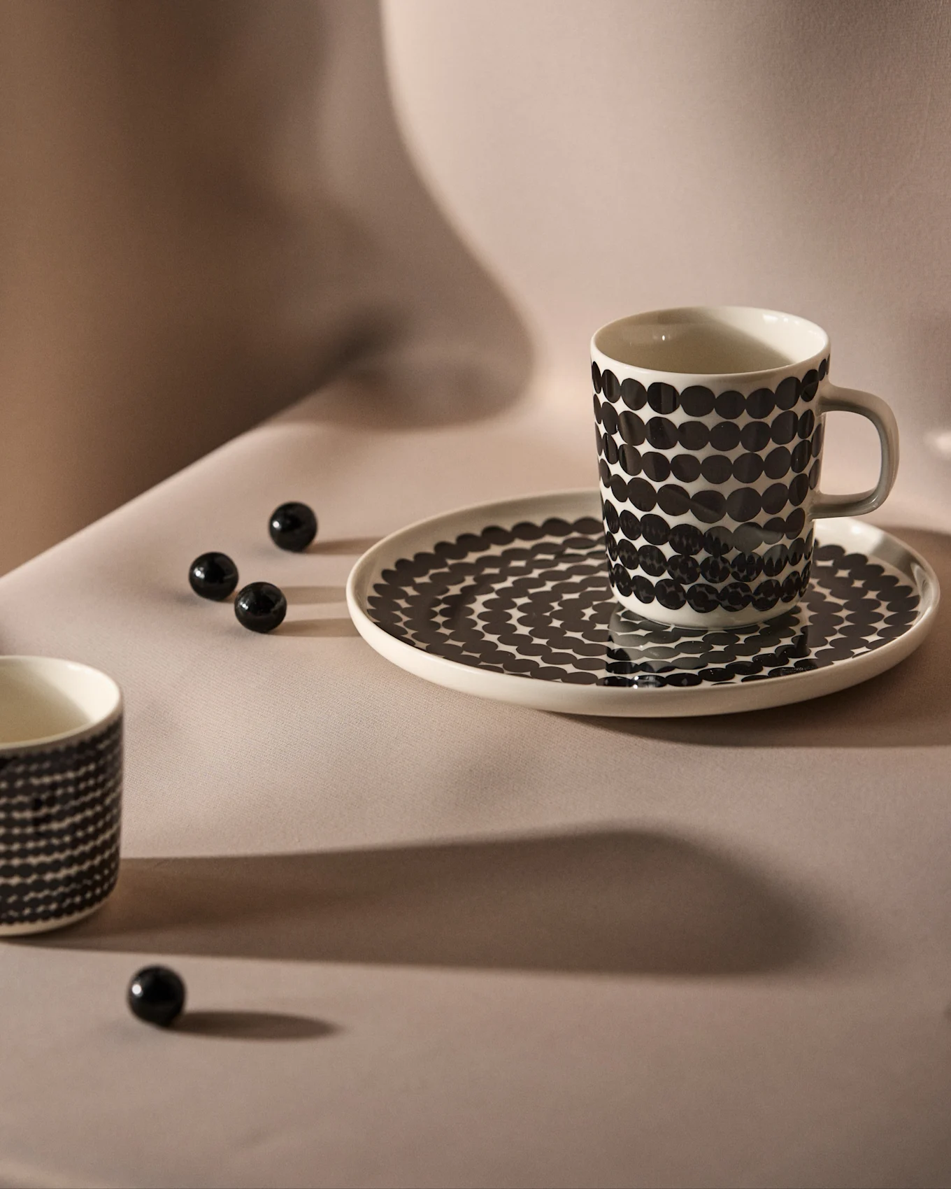 Taza y platillo blancos con estampado de lunares negros sobre fondo neutro, con cuentas decorativas negras dispersas.