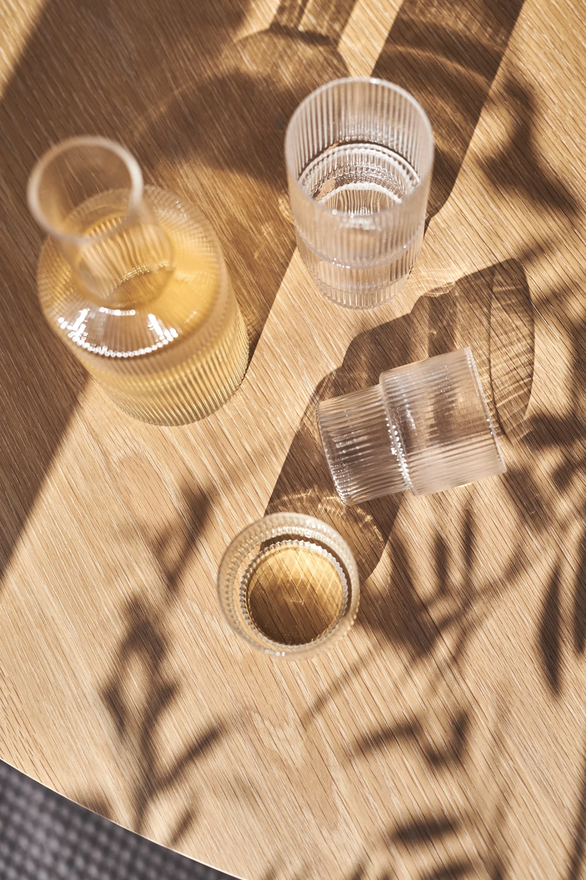 Una botella de vidrio acanalado y dos vasos acanalados, uno con líquido, sobre una mesa de madera soleada con sombras de hojas.