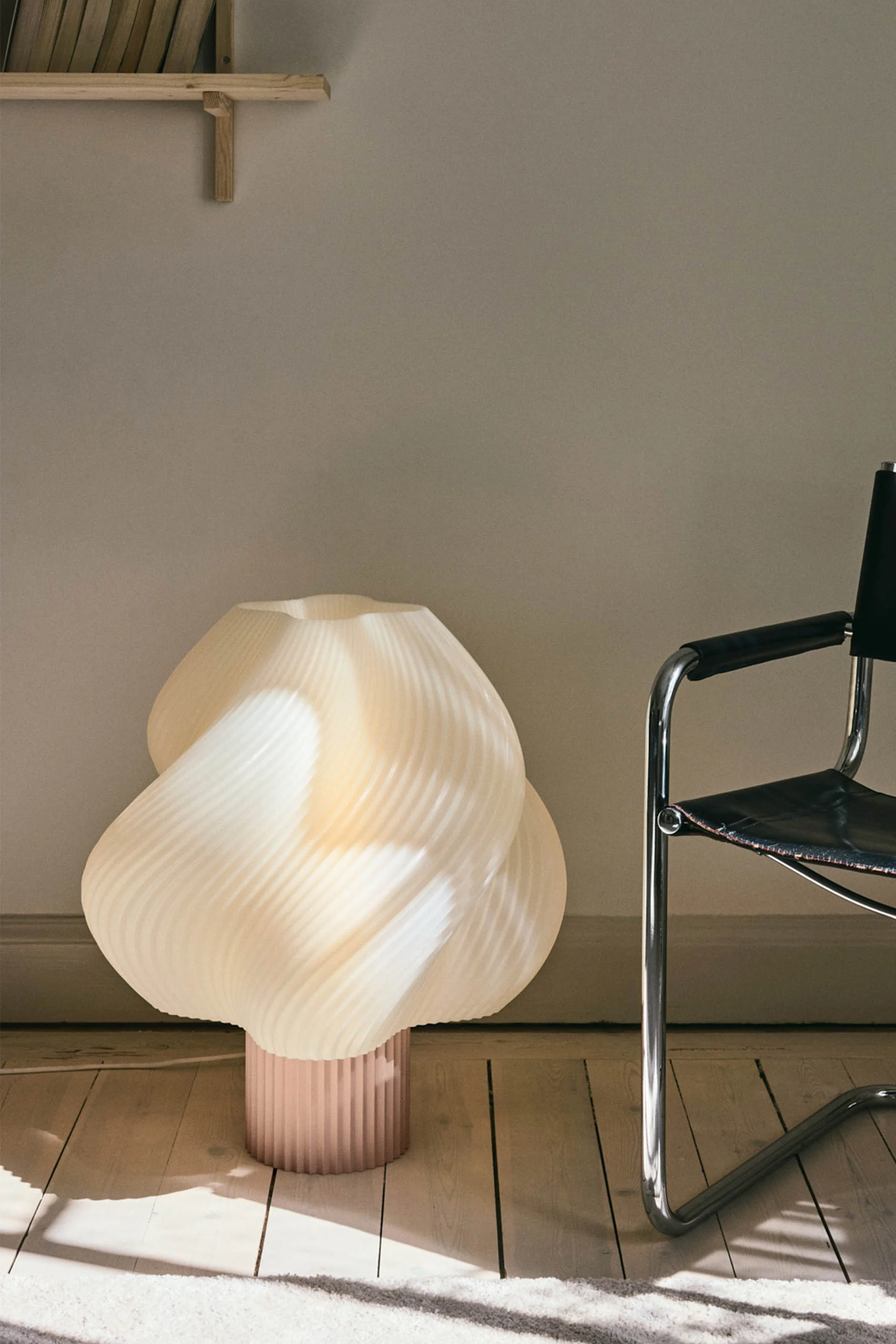 Una lámpara de pie escultural plisada color crema con base rosa se encuentra en un suelo de madera junto a una silla de cuero negro y cromo.