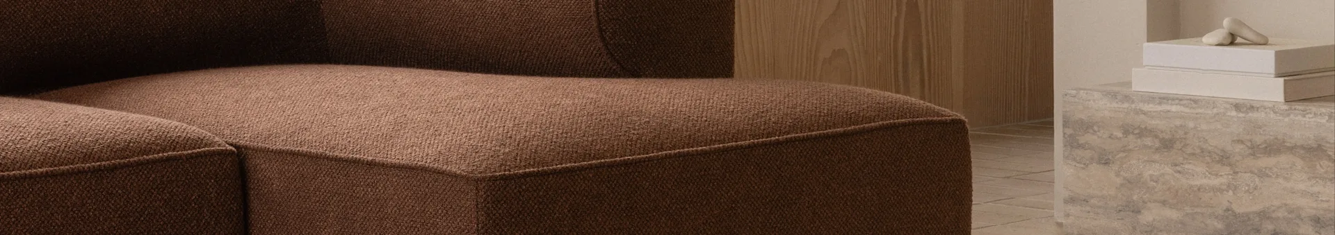 Primer plano de un sofá marrón texturizado, con pared de madera y objetos decorativos sobre hormigón al fondo.