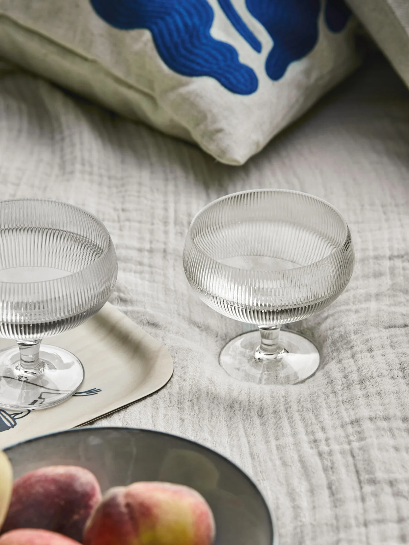 Dos cuencos de postre de vidrio acanalado, un cojín con arte abstracto azul, una bandeja de madera y melocotones sobre tela clara.