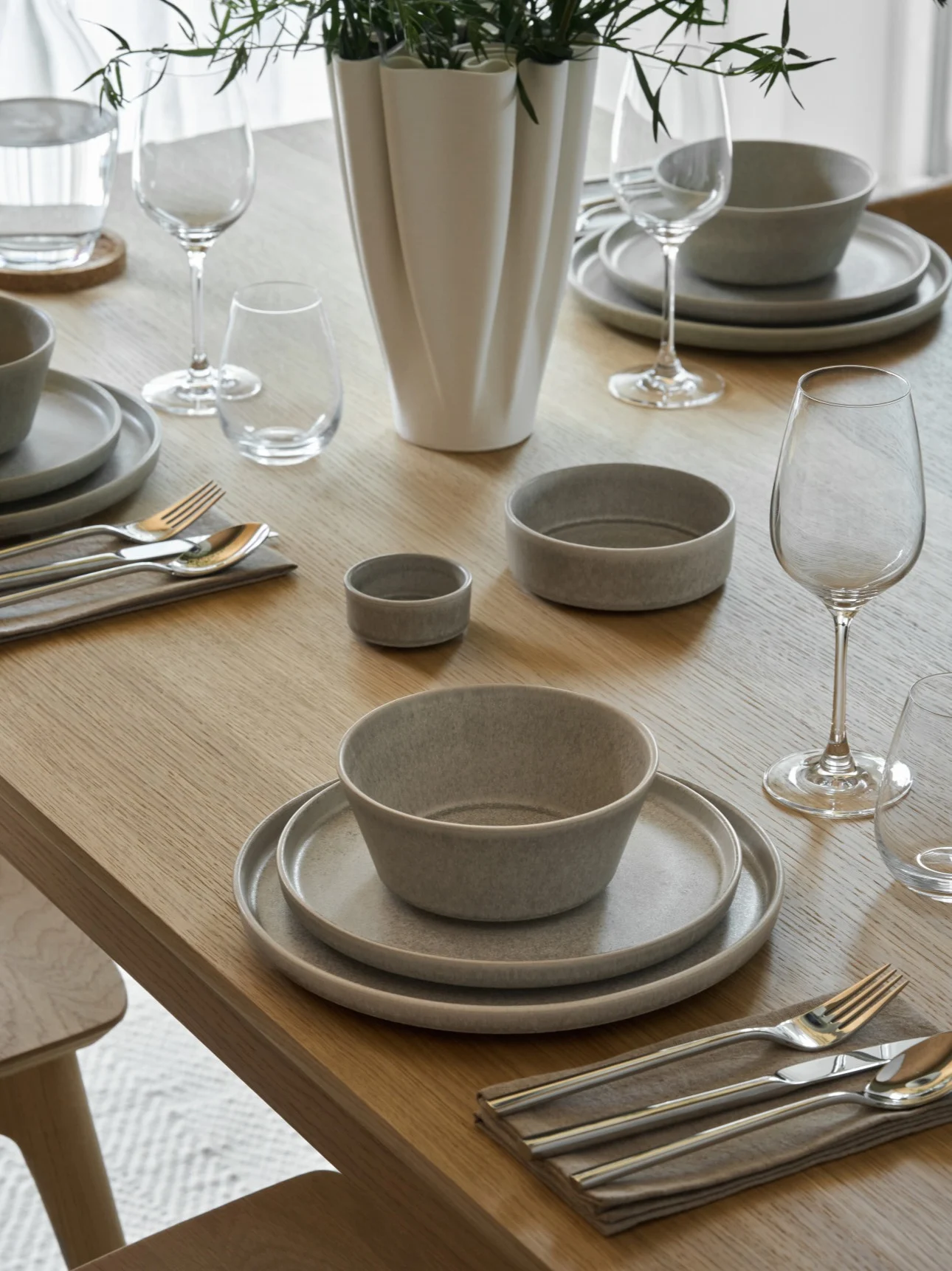 Una mesa de comedor de madera clara puesta con vajilla de cerámica gris, cubiertos de plata, vasos transparentes y un jarrón blanco con follaje verde.