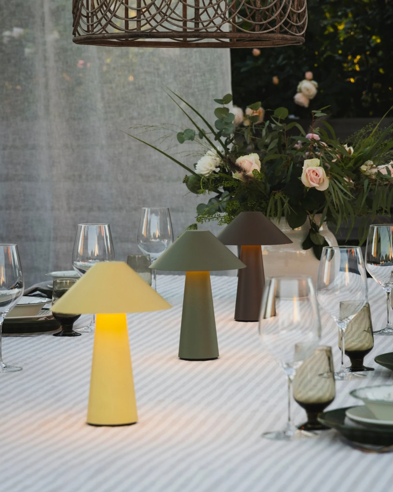 Mesa de comedor exterior con tres lámparas de hongo iluminadas, centro de mesa floral y lámpara colgante de mimbre.
