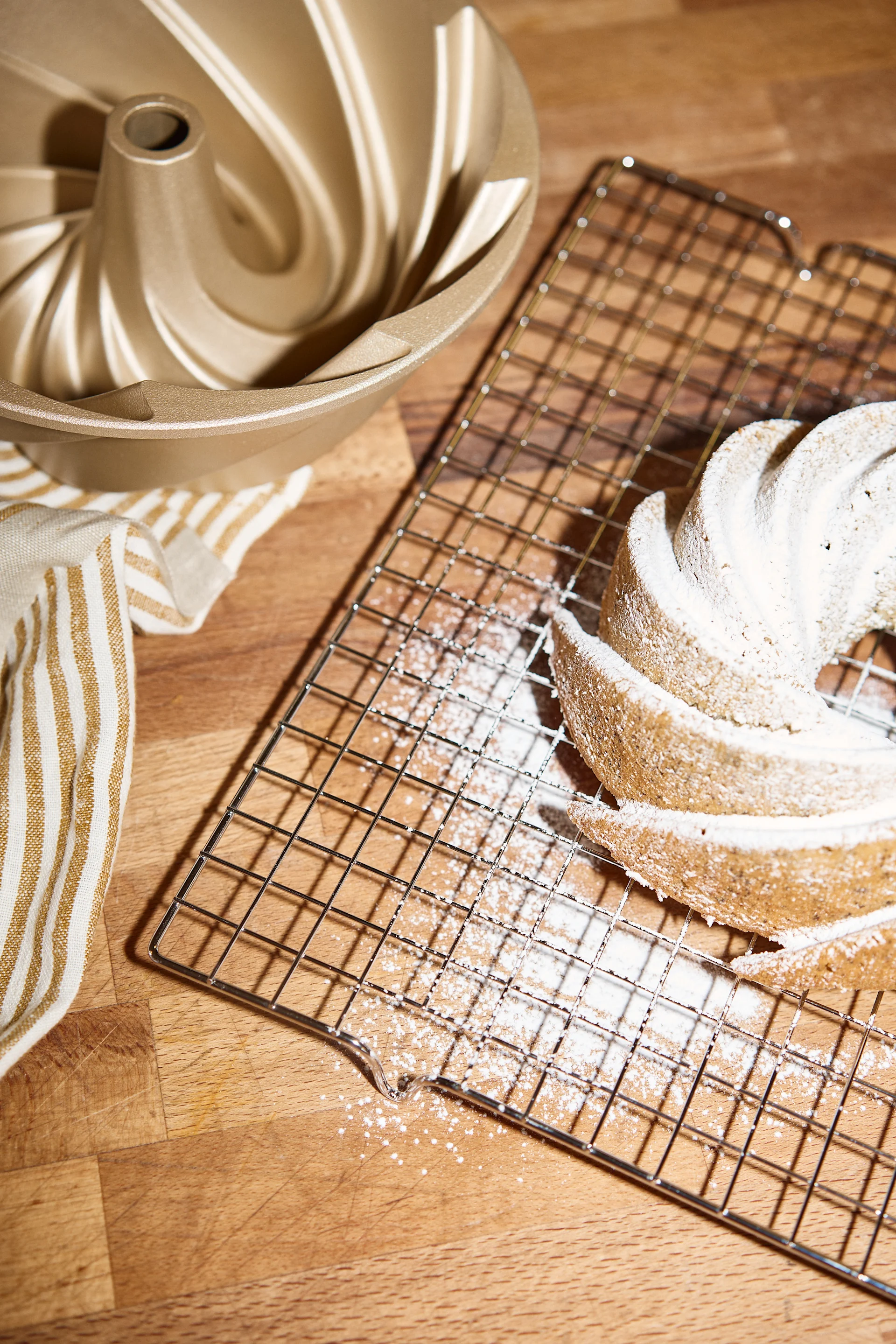 Un molde bundt dorado, una toalla a rayas y un bizcocho bundt espolvoreado con azúcar glas en una rejilla.