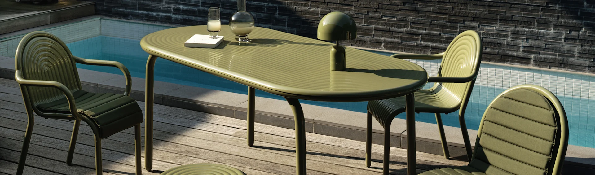 Mesa y sillas de comedor verdes acanaladas al aire libre en una terraza de madera junto a una piscina. Una lámpara, jarra, vaso y libro están sobre la mesa.
