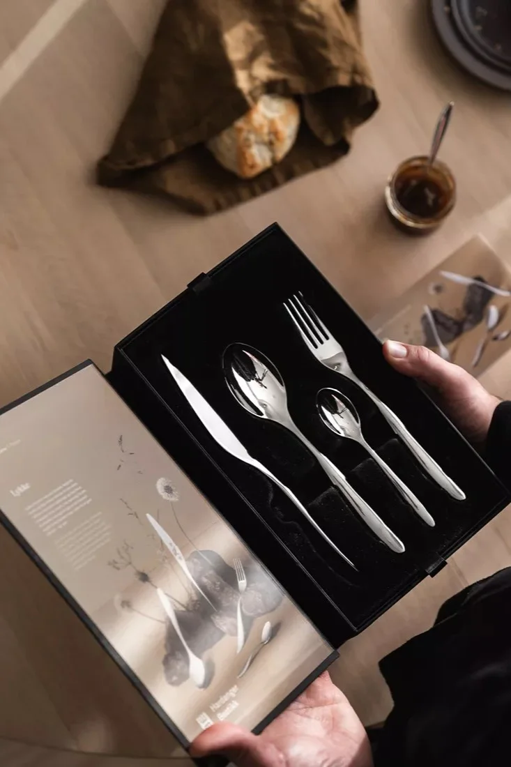 Manos sostienen caja negra abierta con un juego de cubiertos de plata brillantes: cuchillo, cuchara grande, cuchara pequeña y tenedor. Pan y mermelada en mesa de madera.