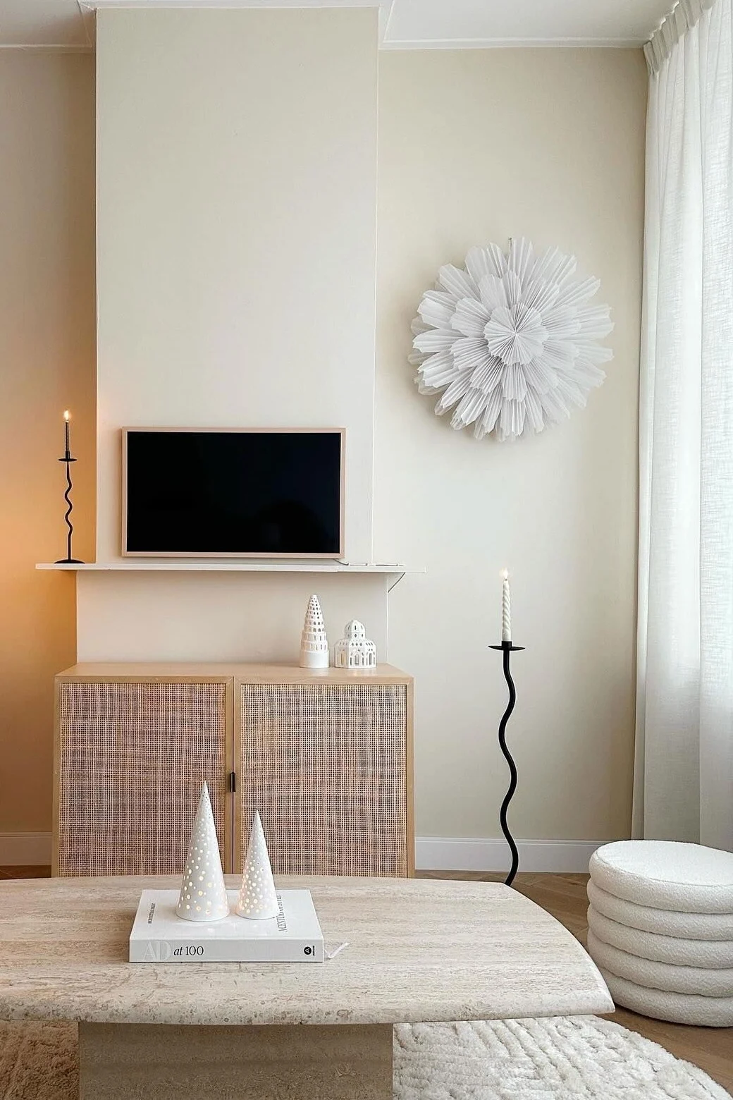 Salón neutro con alfombra texturizada, mesa de travertino, mueble de ratán con TV, lámpara de pie y adorno de pared plisado.