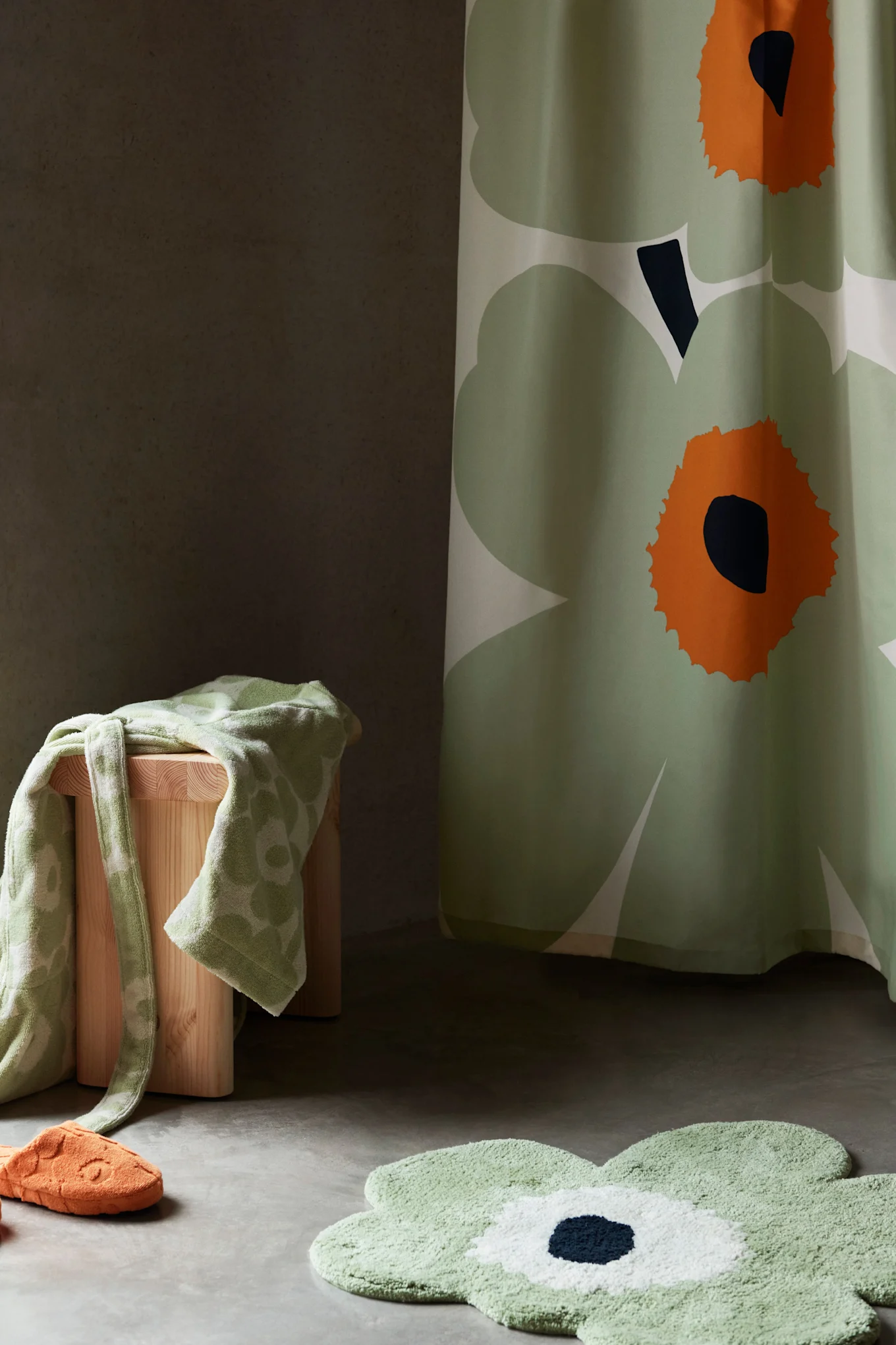 Decoración de baño con una cortina de ducha verde con motivos florales, una alfombra de baño, una toalla sobre un taburete y una zapatilla naranja.
