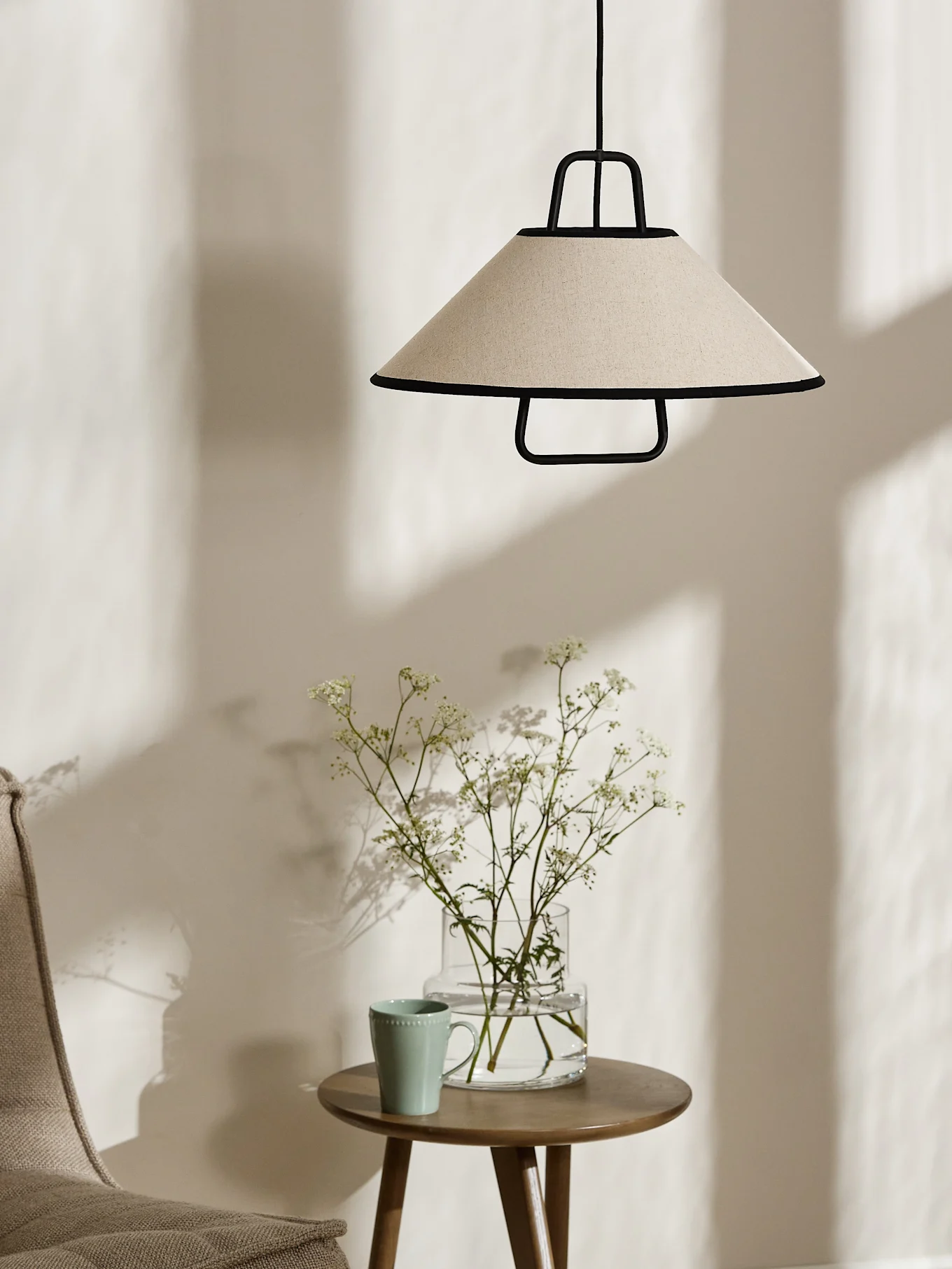 Una lámpara colgante cónica color crema con ribete negro cuelga sobre una mesa auxiliar de madera con un jarrón de flores blancas y una taza verde claro.