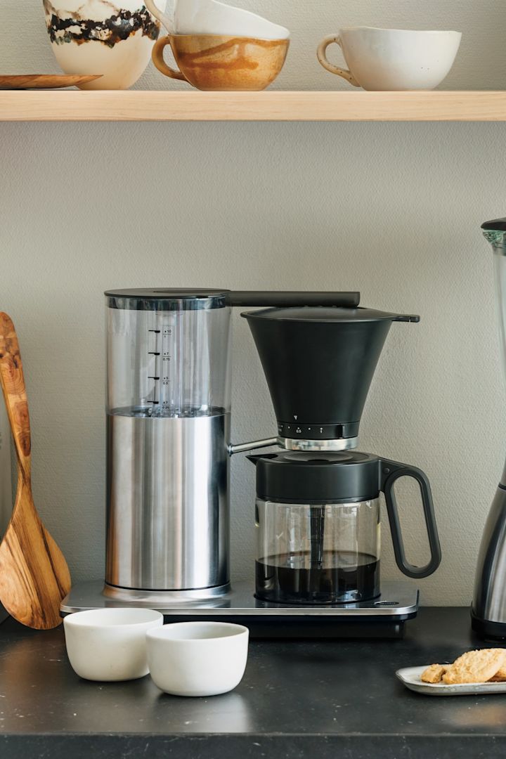 También necesitas los electrodomésticos adecuados para tu rincón del café en casa. Aquí puedes ver una cafetera de filtro clásica de Wilfa en un aparador de cocina, con una estantería con cuencos y una tostadora encima.
