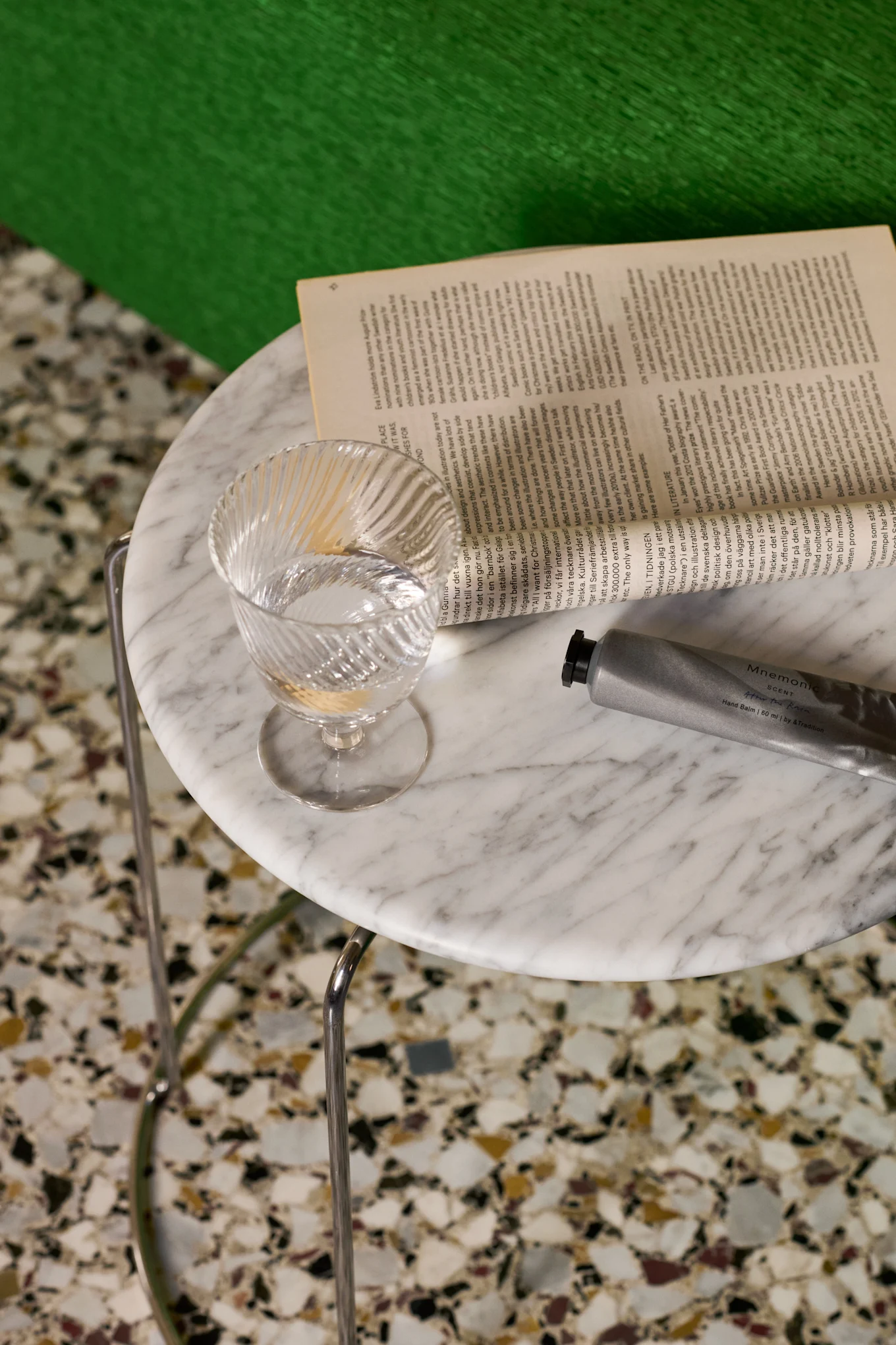 Un vaso de agua transparente y estriado, un libro abierto y un tubo de bálsamo de manos plateado sobre una mesa de mármol.