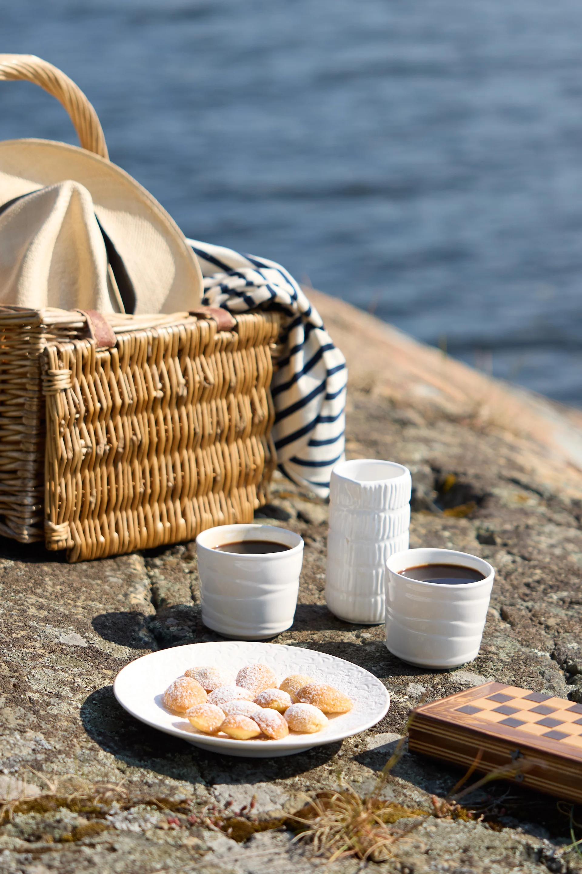 Un picnic sobre rocas junto al mar, con una cesta de mimbre con sombrero y manta a rayas, dos tazas de café, pasteles y un tablero de ajedrez cerrado.