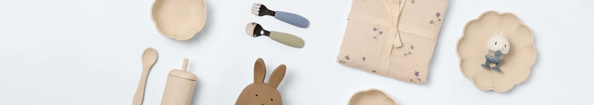 Artículos para bebé sobre blanco: dos cuencos crema, cubiertos azules y verdes, tela floral, juguete de conejo, cuchara de madera, taza beige y figura en plato festoneado.